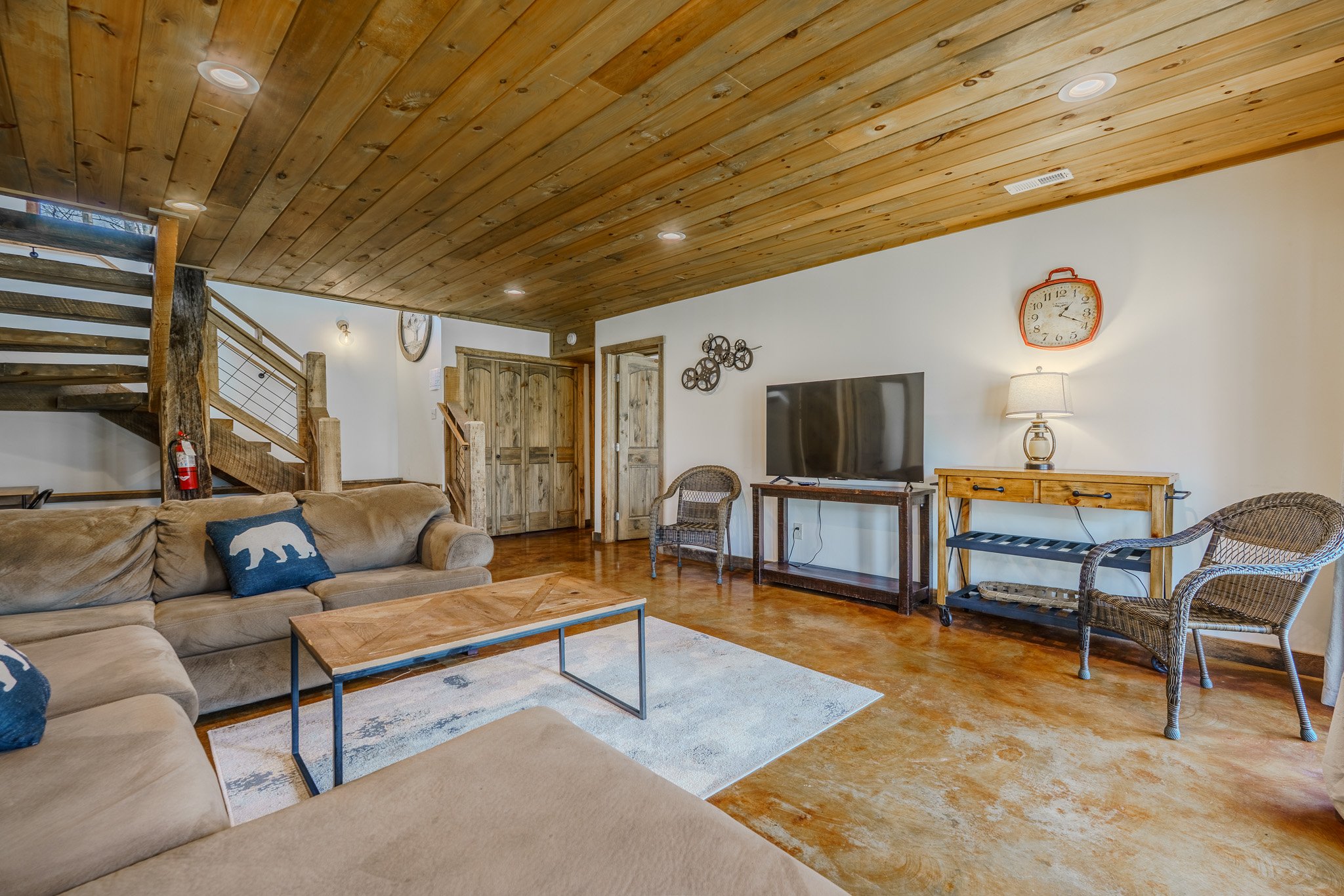 Living room with wooden ceiling and floor, beige sectional sofa with bear-themed pillows, coffee table, TV on wooden stand, wicker chairs, wooden console table with lamp, rustic wooden doors, wall clock, staircase with wood and metal railing, wall de