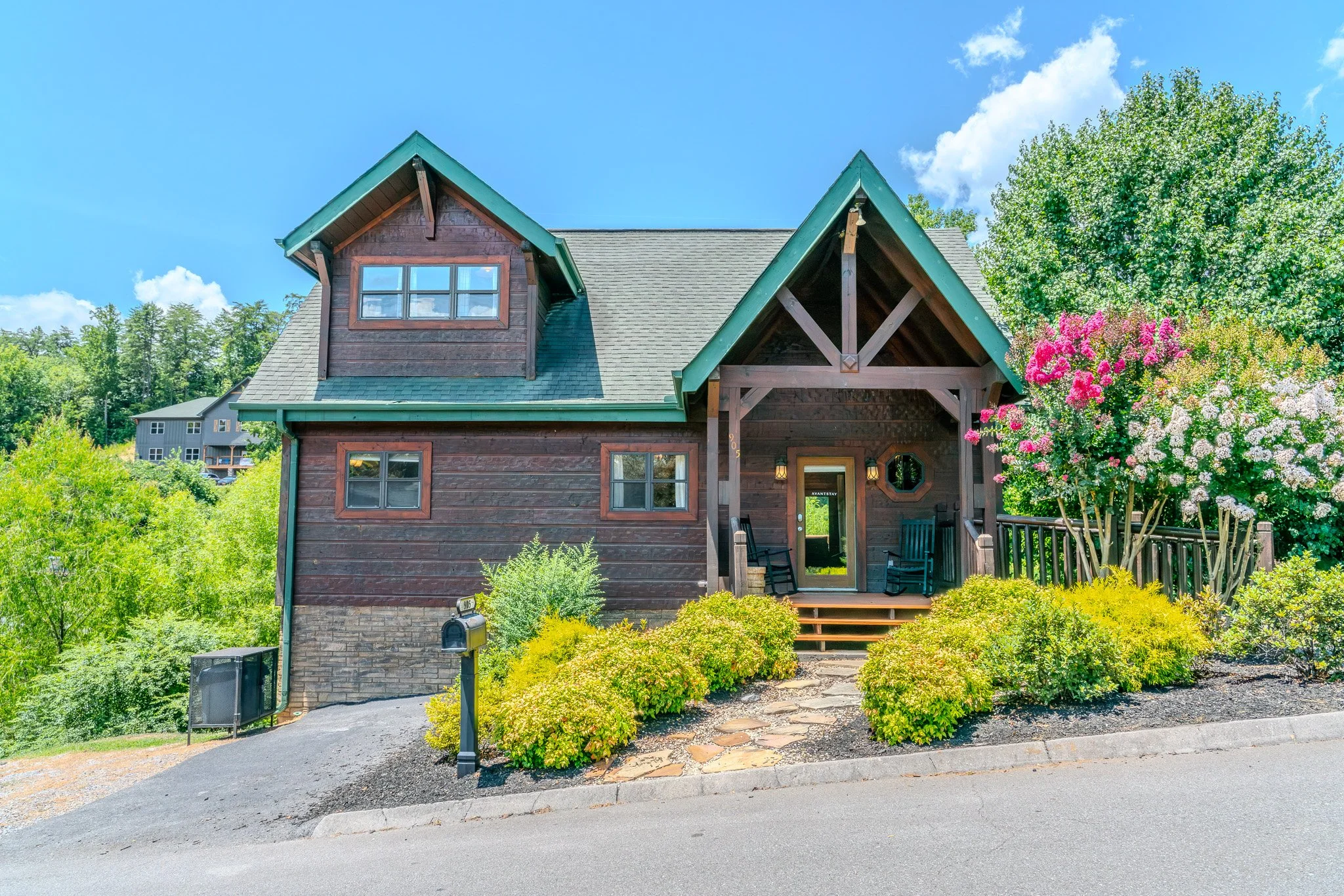 Wooden house with a green roof, front porch with black rocking chairs, surrounded by colorful bushes and pink and white flowering bushes, blue sky with clouds