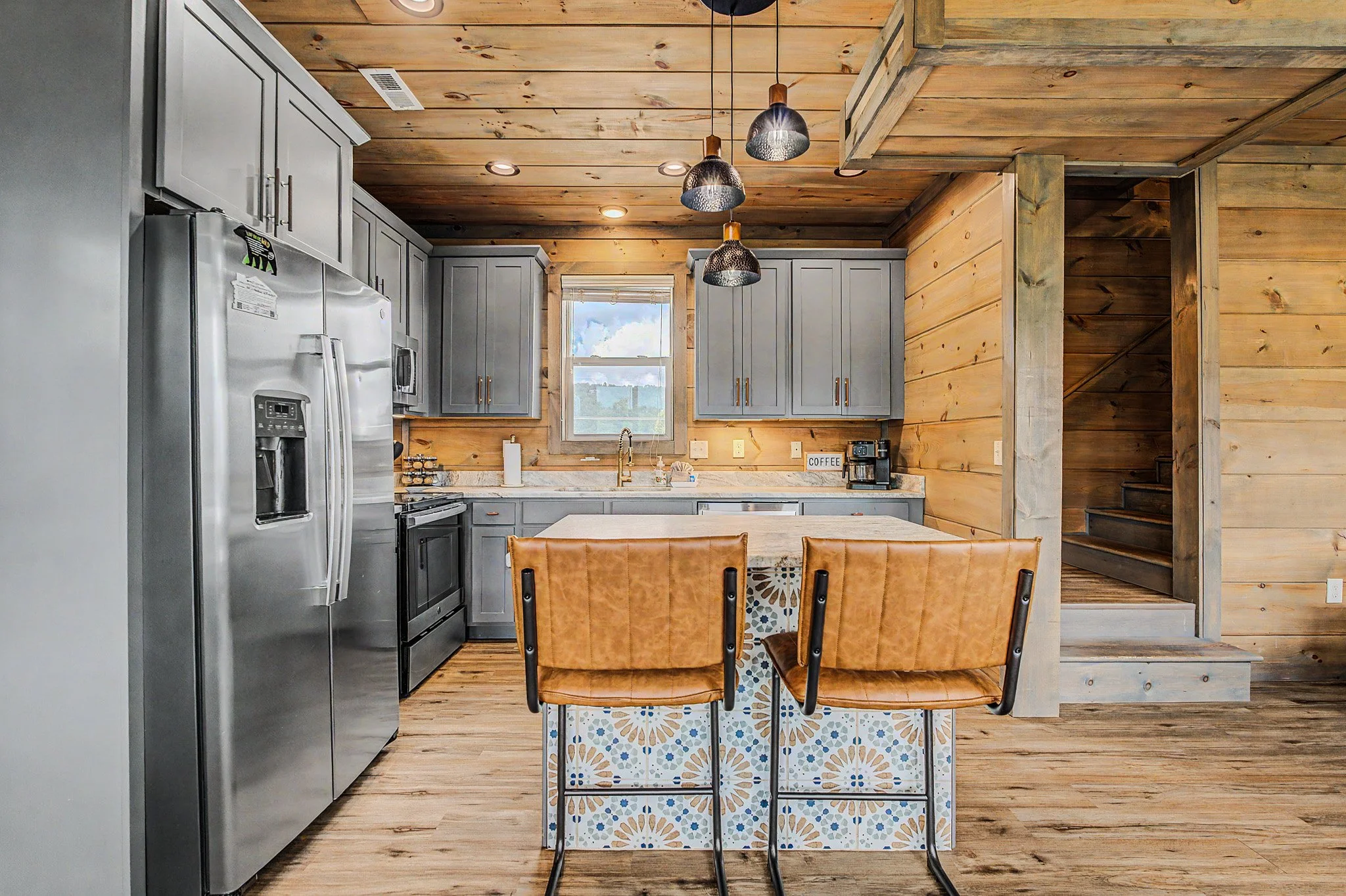 A kitchen with light gray cabinets, a stainless steel refrigerator, a window above the sink, wooden walls and ceiling, three pendant lights, a white countertop with a coffee machine, and a decorative tiled kitchen island with two leather chairs in th