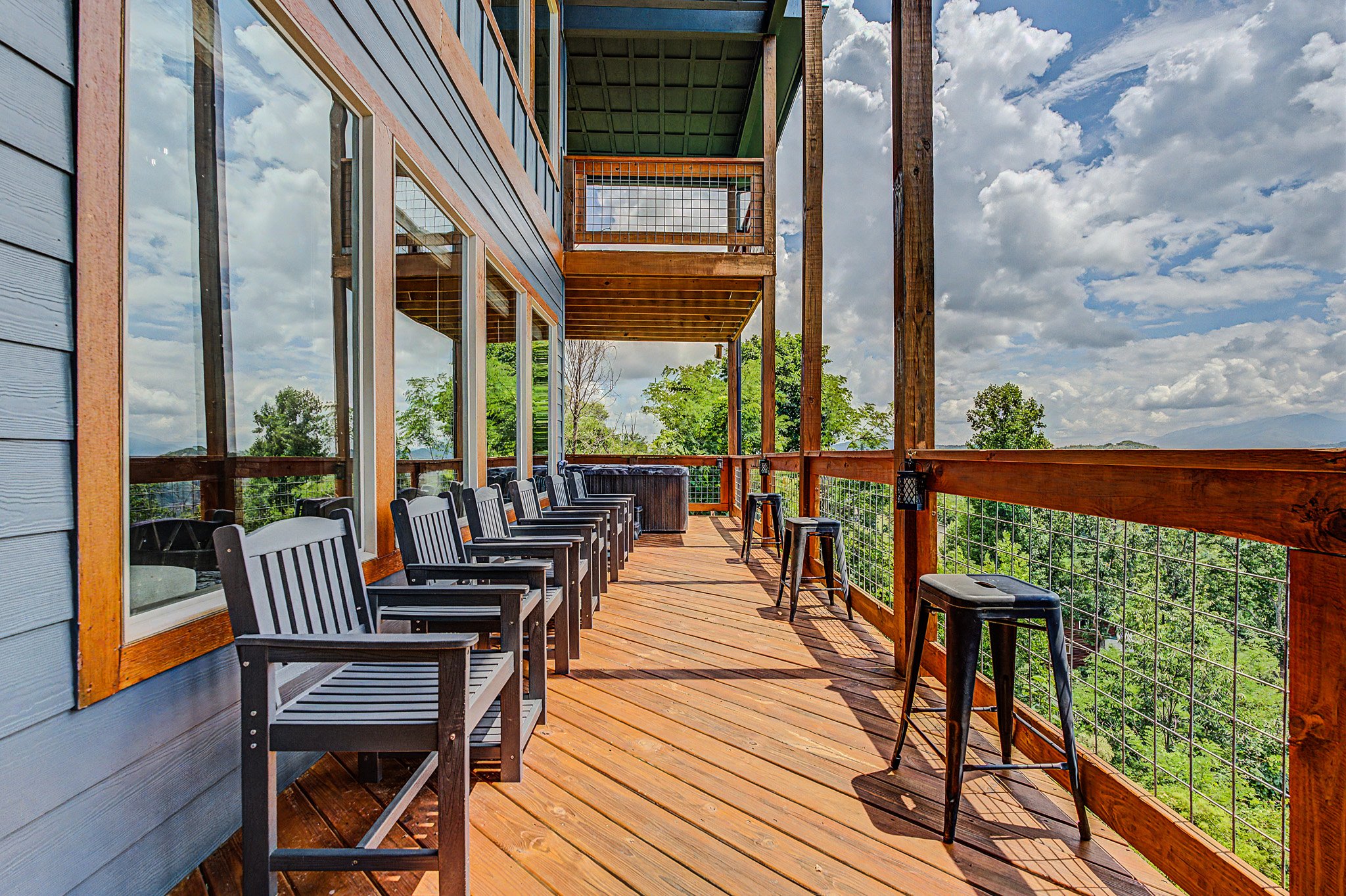 A spacious wooden balcony or deck attached to a blue house with large windows, overlooking green trees and a partly cloudy sky, with chairs lined up along the railing.