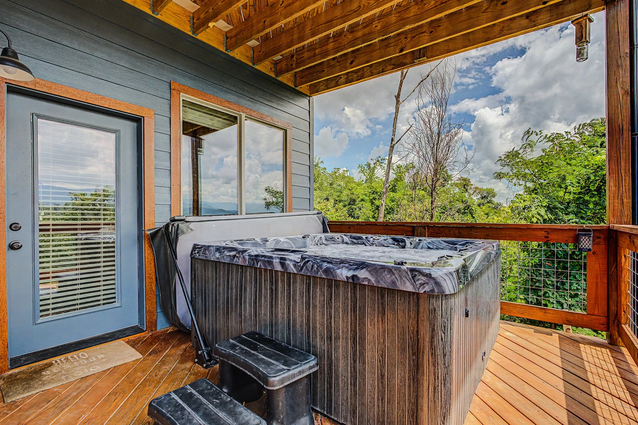 On a wooden porch, a hot tub with a marble-patterned exterior, a black stool, and steps leading into the hot tub. There is a door with a window and blinds, a window beside it, and trees and cloudy sky in the background.