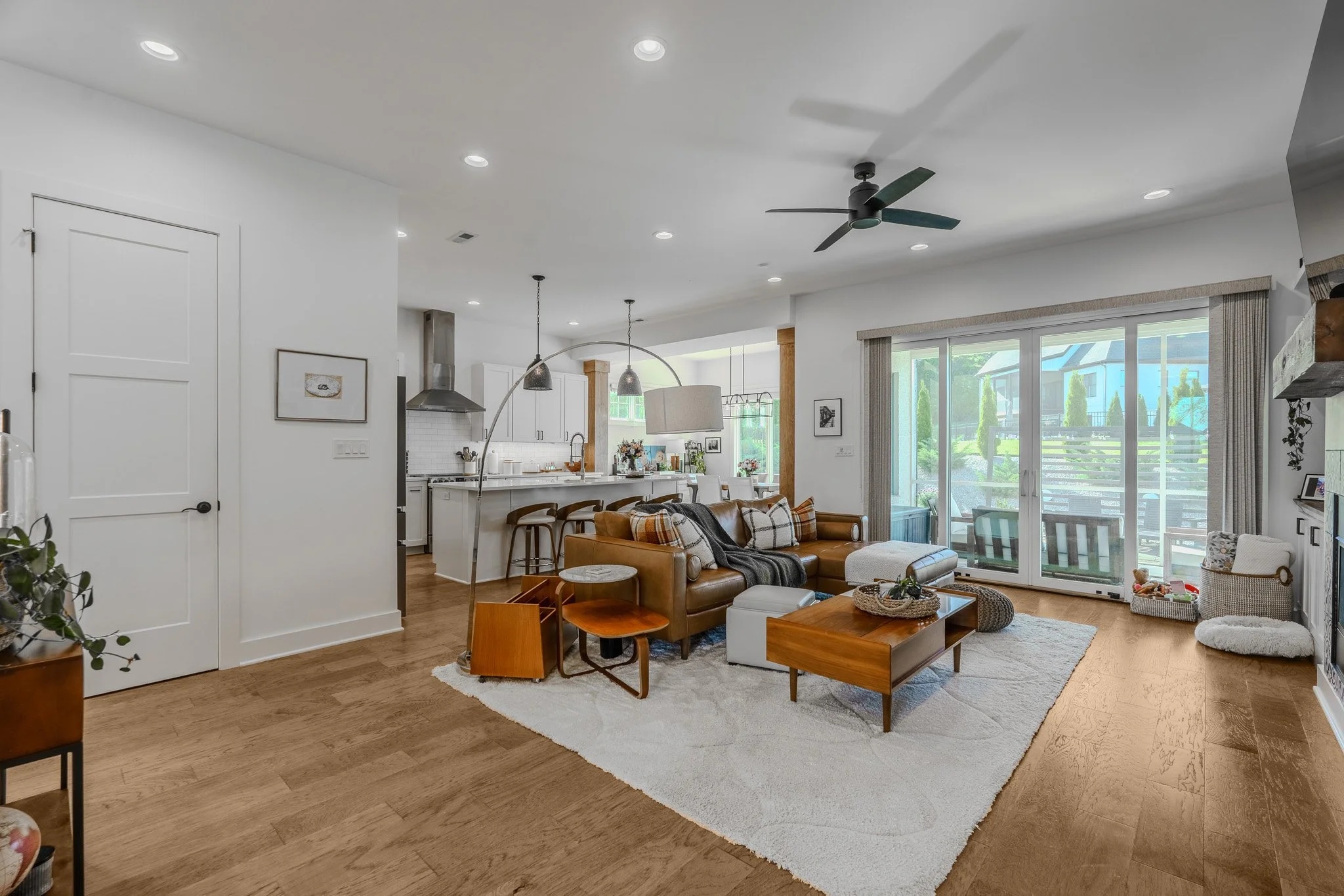 Open concept living room and kitchen with sliding glass door leading to outdoor patio, brown leather sofa, wooden coffee table, white rug, and large windows providing natural light.