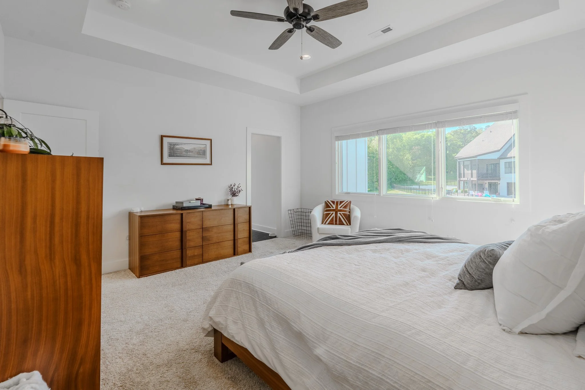 Bright bedroom with large window, white walls, beige carpet, bed with white linens, wooden dresser, white armchair with Union Jack pillow, ceiling fan, framed picture on wall.