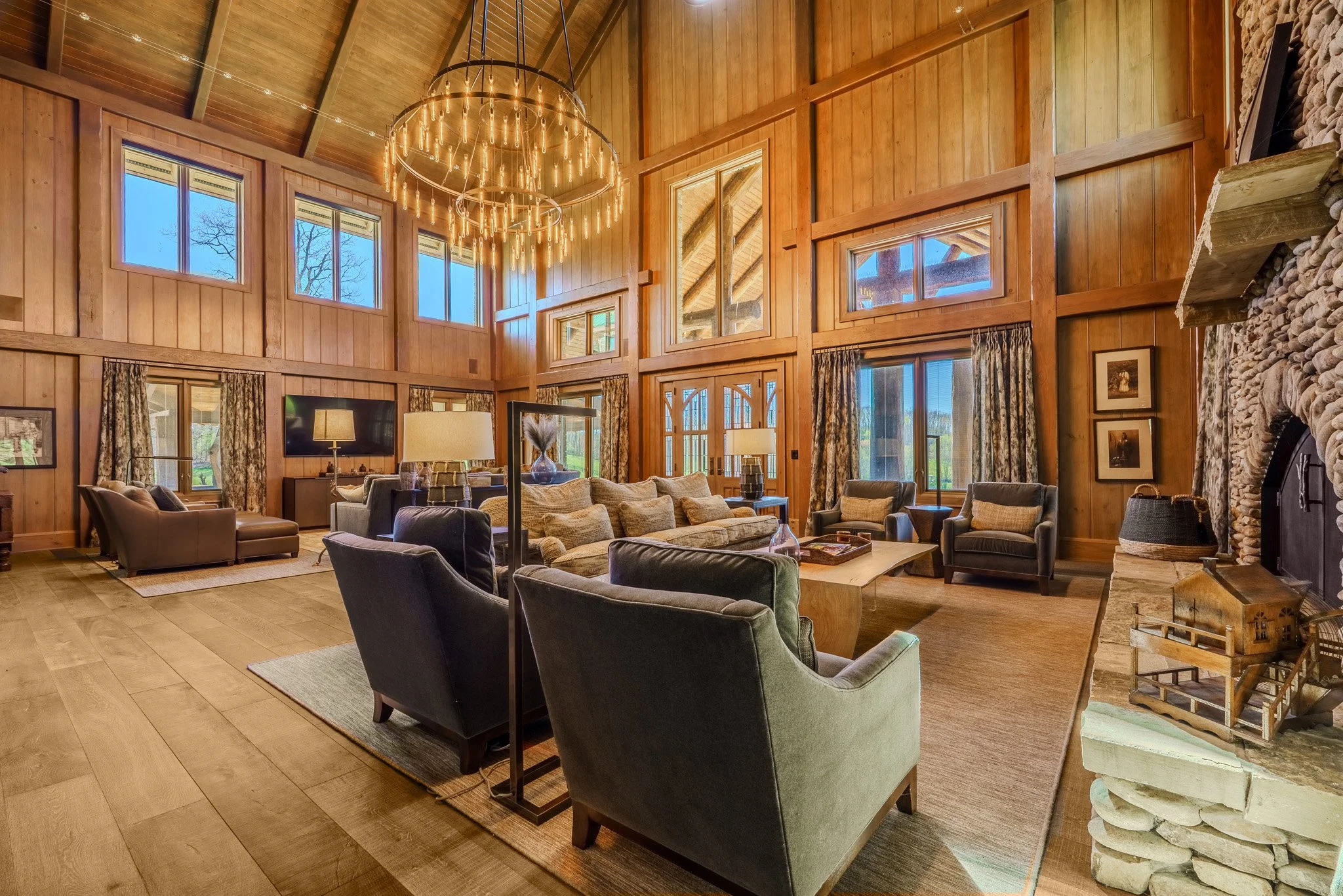 Loft-style living room with high ceilings, large windows, wooden panel walls, chandelier, fireplace, and various chairs and sofas.