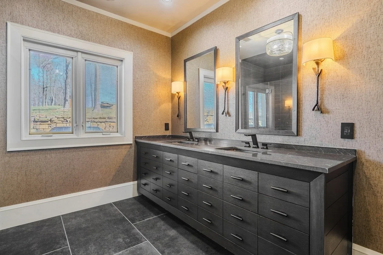 A modern bathroom vanity with two sinks, two mirrors, side wall sconces, a large window with blinds, dark gray cabinetry, and a gray tiled floor.