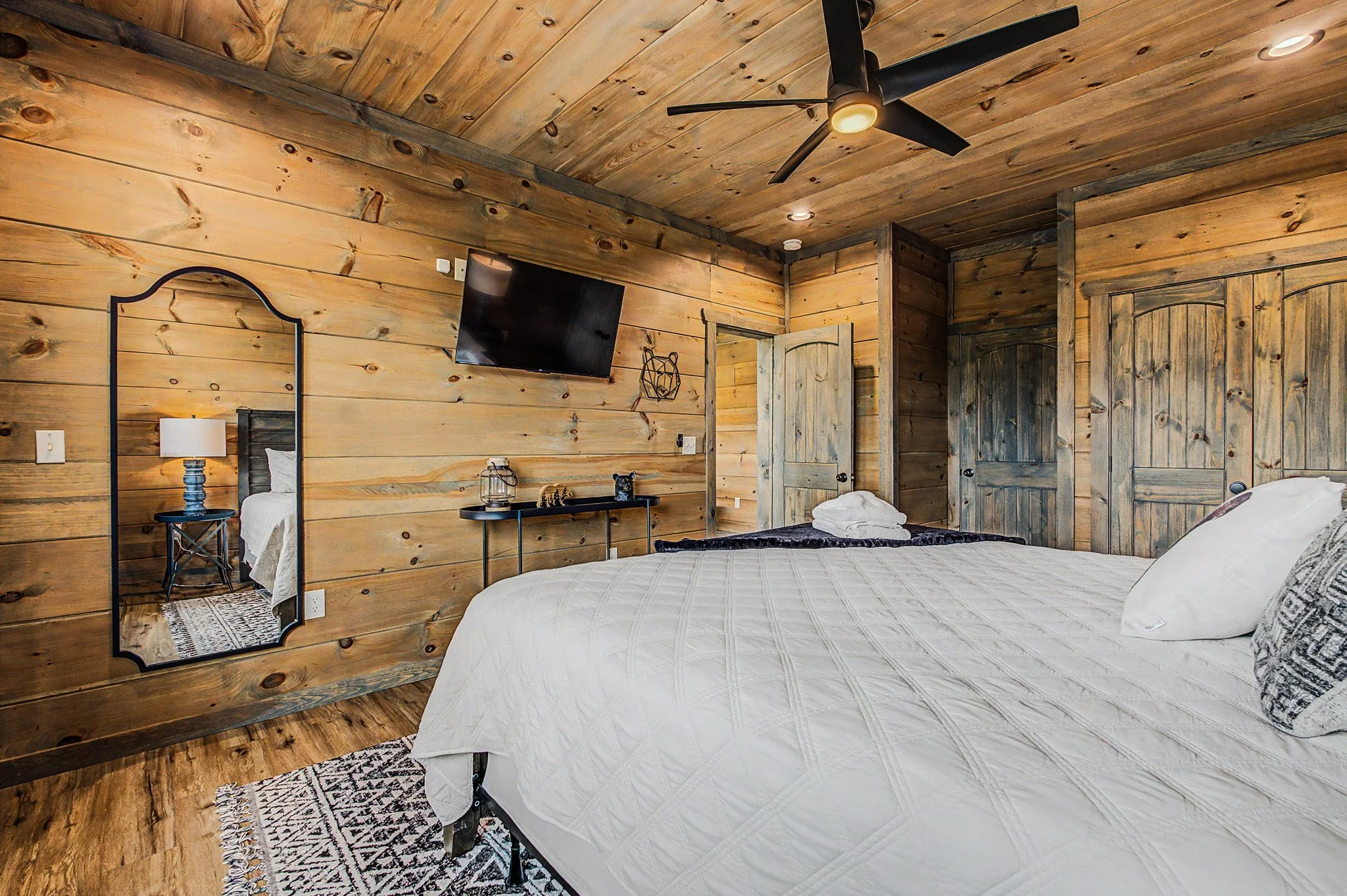 A rustic bedroom with wood-paneled walls and ceiling, a large bed with white bedding, a mirror, a TV mounted on the wall, a nightstand with a lamp, and a wooden door.
