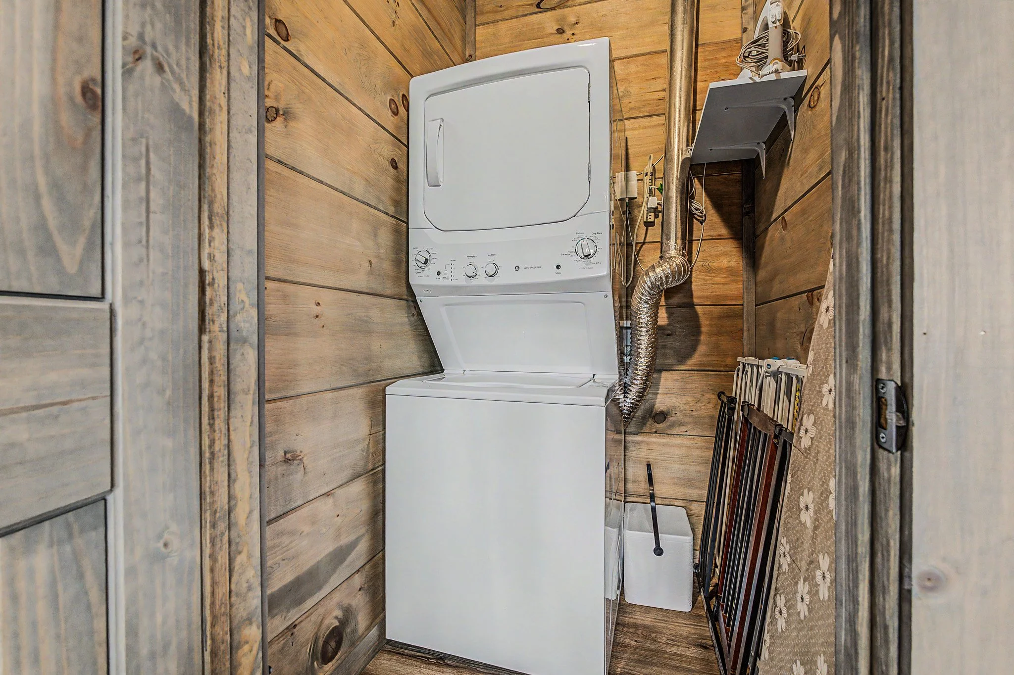 The image shows a small laundry area with a white top-loading dryer, wooden wall paneling, some electrical cords, and metal ductwork. There are some cleaning supplies and a ladder or similar equipment leaning against the wall on the right side.