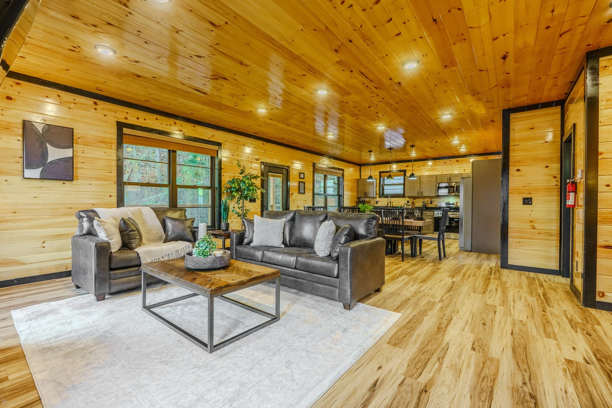 A living room and kitchen area with wood-paneled walls and ceiling, featuring leather sofas, a coffee table, large windows, and a dining table, with a hardwood floor.