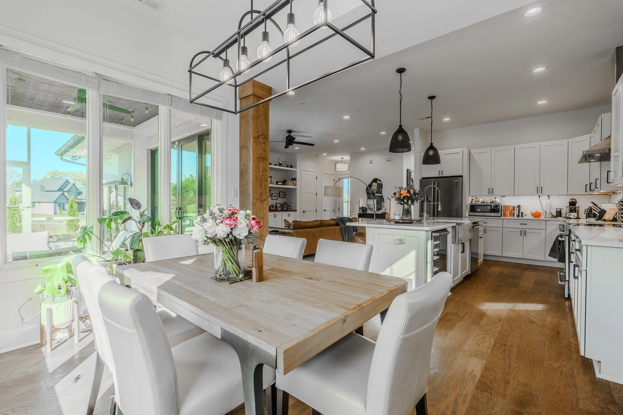 Open-concept kitchen and dining area with white cabinets, a wooden dining table, white chairs, black pendant lights, and large sliding glass doors leading to an outdoor patio with a garden.