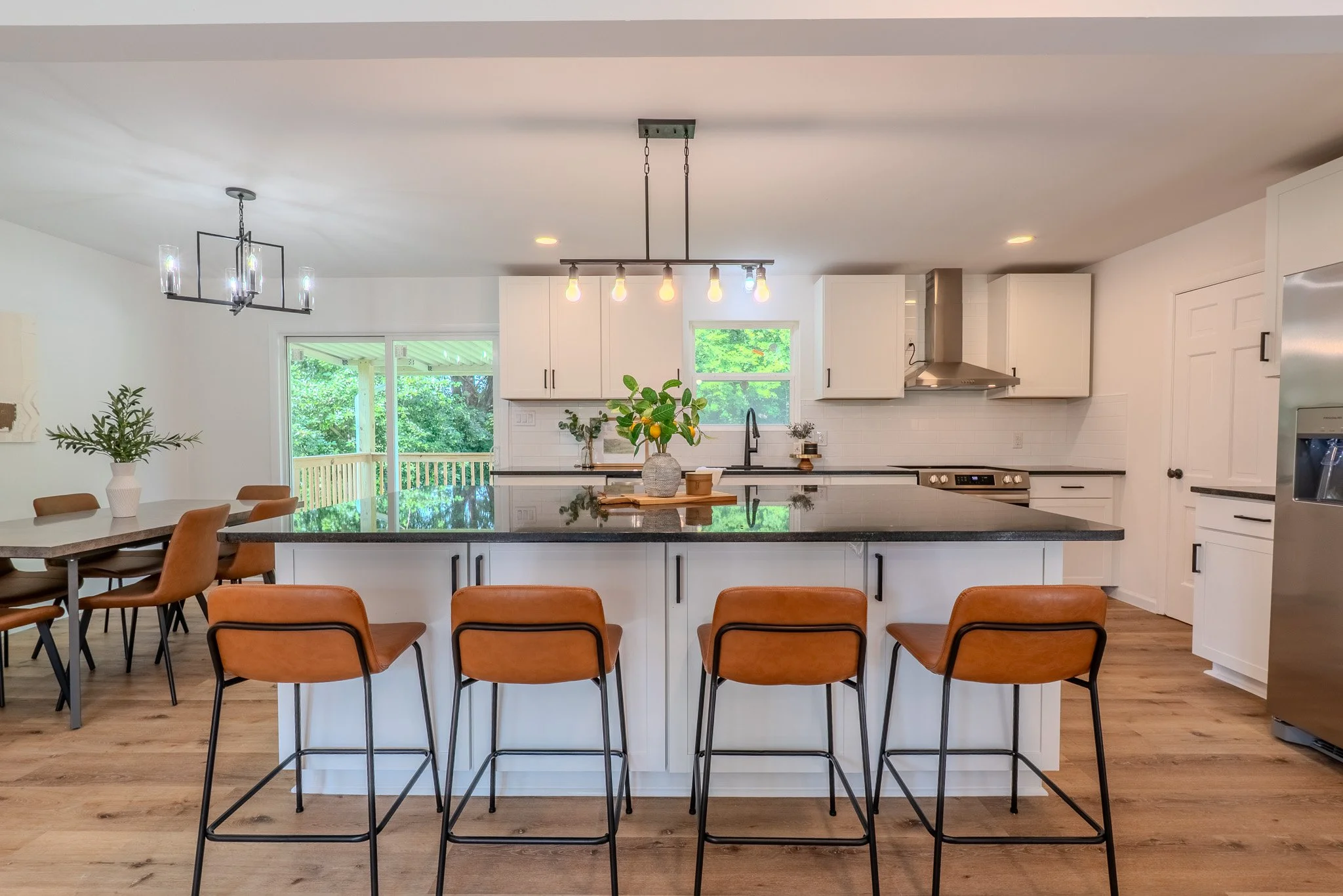 Modern kitchen with white cabinets, black countertops, a kitchen island with bar stools, wooden dining table with chairs, stainless steel appliances, and pendant lighting, overlooking a green outdoor area.