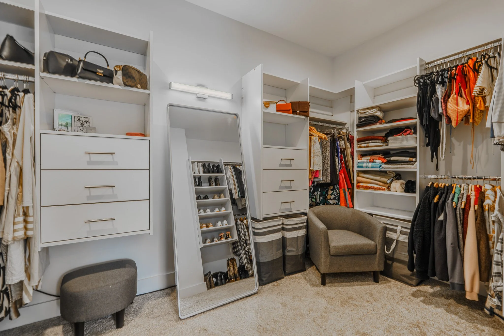 Organized walk-in closet with shelves, hanging clothes, shoes, handbags, chair, and a mirror.