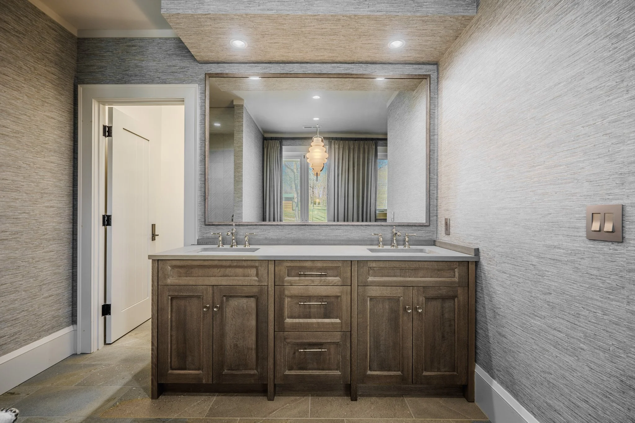 Bathroom vanity with a large mirror, two sinks, wooden cabinet, and gray textured walls.
