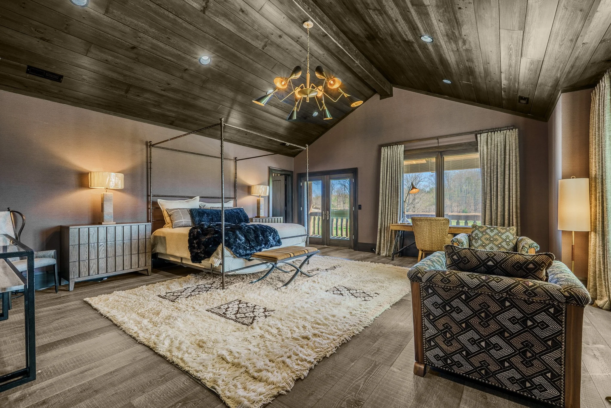 Spacious living room with wooden ceiling, large window, glass door leading outside, overstuffed sofa, patterned armchair, side tables with lamps, and a four-poster bed in the background, decorated with a modern chandelier.