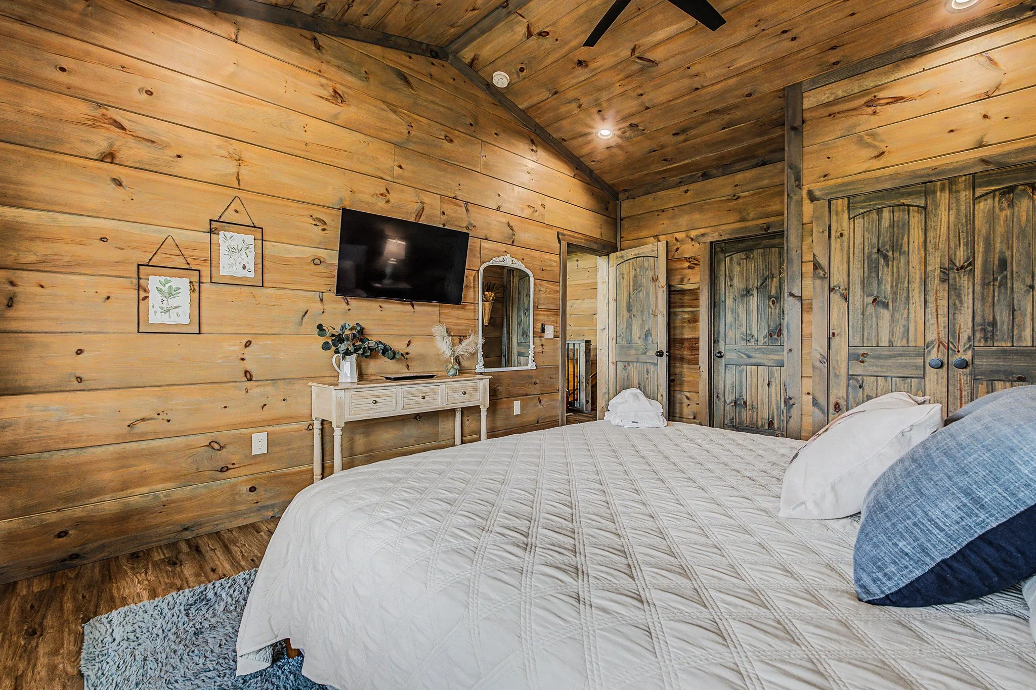 A rustic bedroom with wood-paneled walls and ceiling, featuring a mounted flat-screen TV, a white dresser with decorative items, and a bed with white bedding and pillows. There are two wooden closet doors and a mirror on the wall.