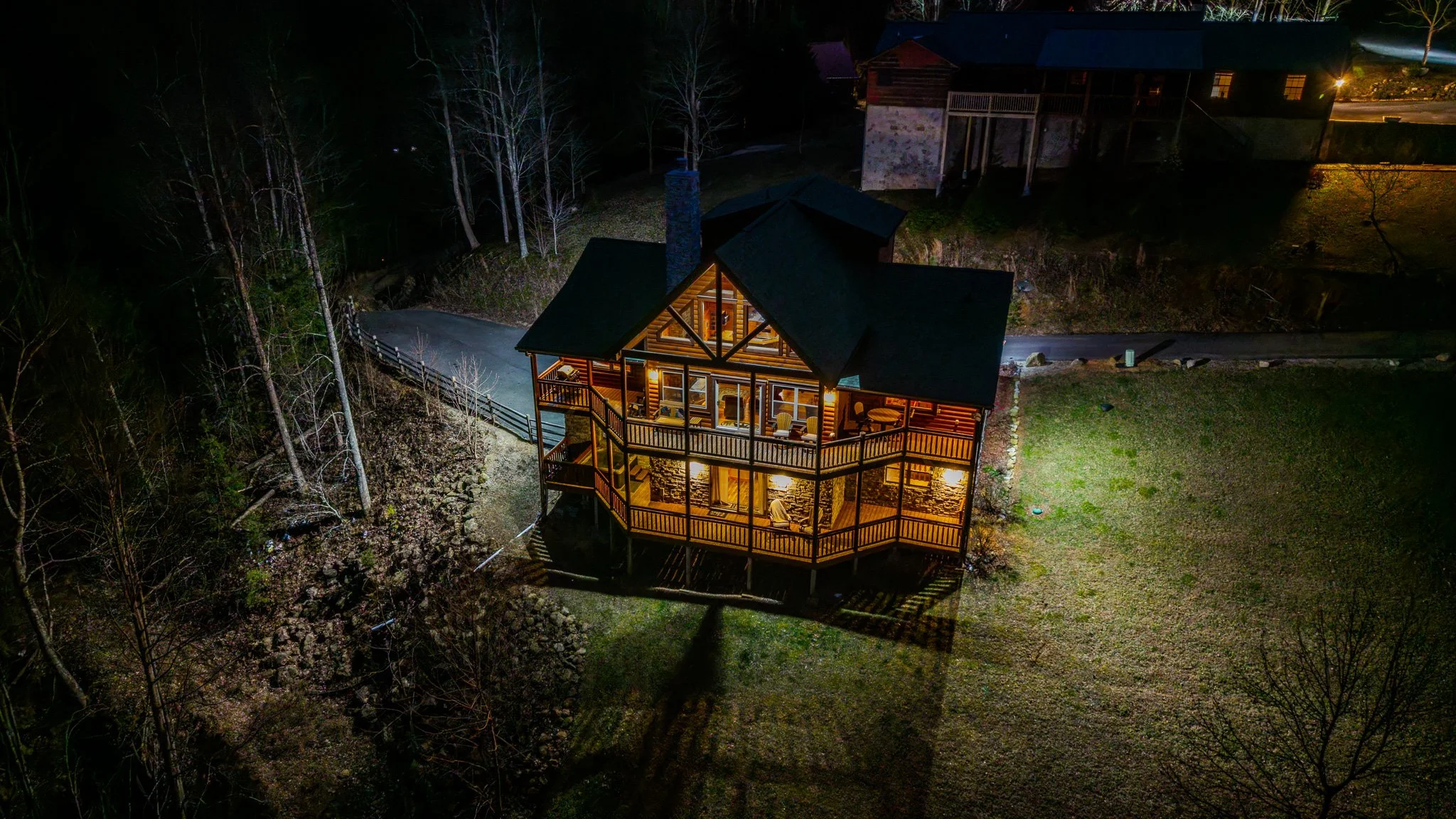 A two-story wooden house with a large deck, lit up from within, situated on a hillside at night. Surrounding features include trees, a small road, and a grassy yard with sparse trees.