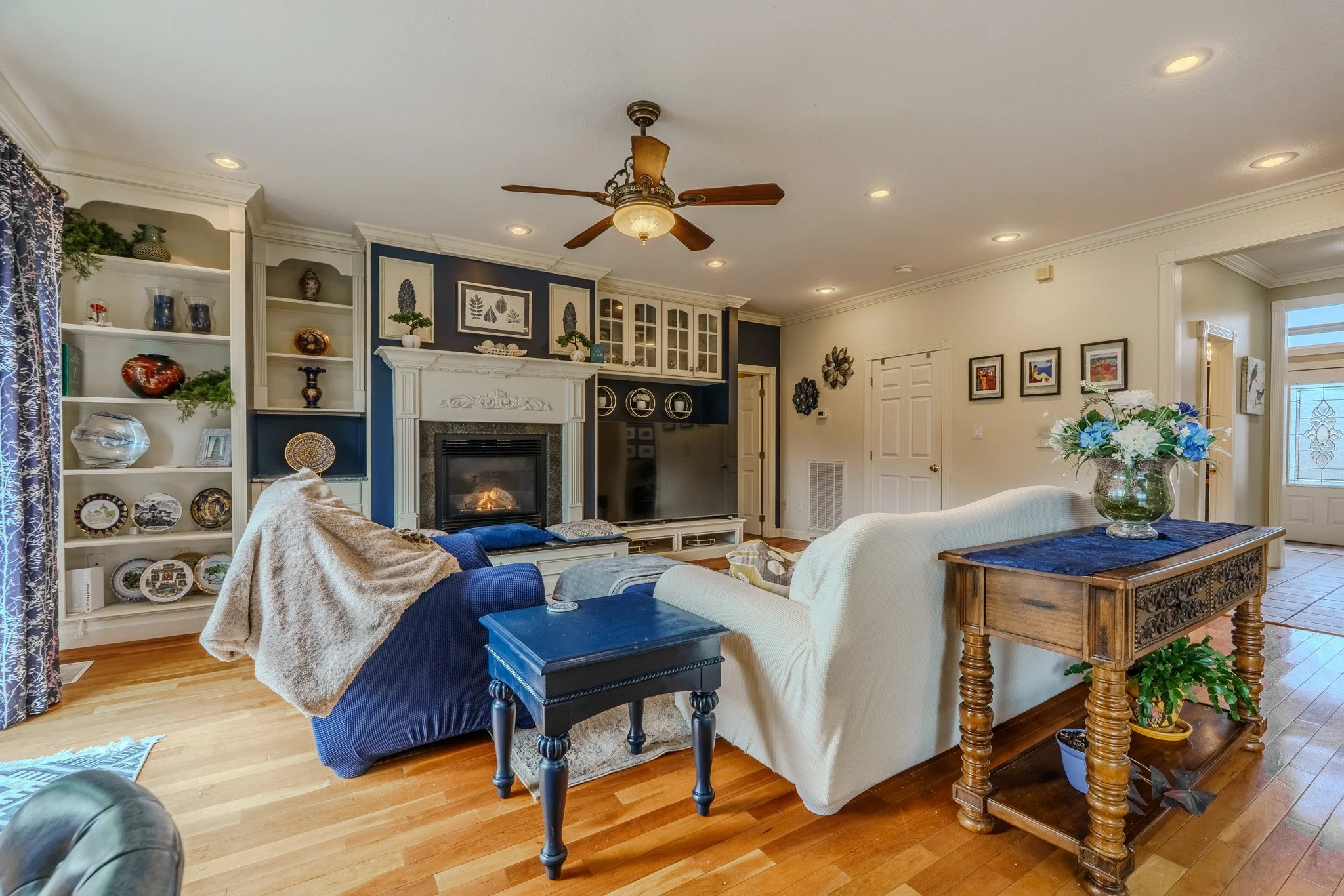 Living room with white and blue walls, hardwood flooring, a white fireplace, built-in shelves with decorative items, a ceiling fan, a white sofa, a blue armchair with a blanket, a television, and a wooden console table with a vase of flowers.