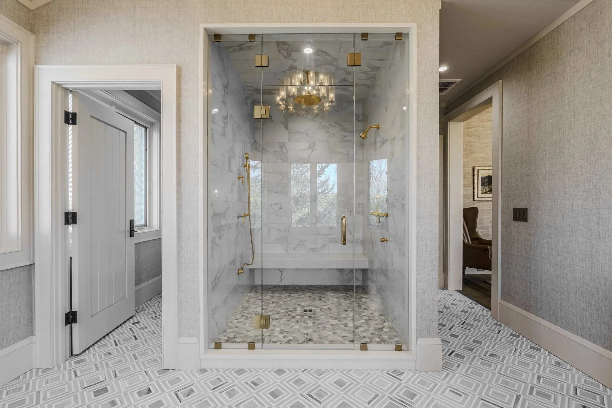A walk-in shower with glass doors and gold fixtures, including two showerheads, a handheld shower, and multiple body sprays, inside a bathroom with patterned tile flooring and light-colored textured walls.