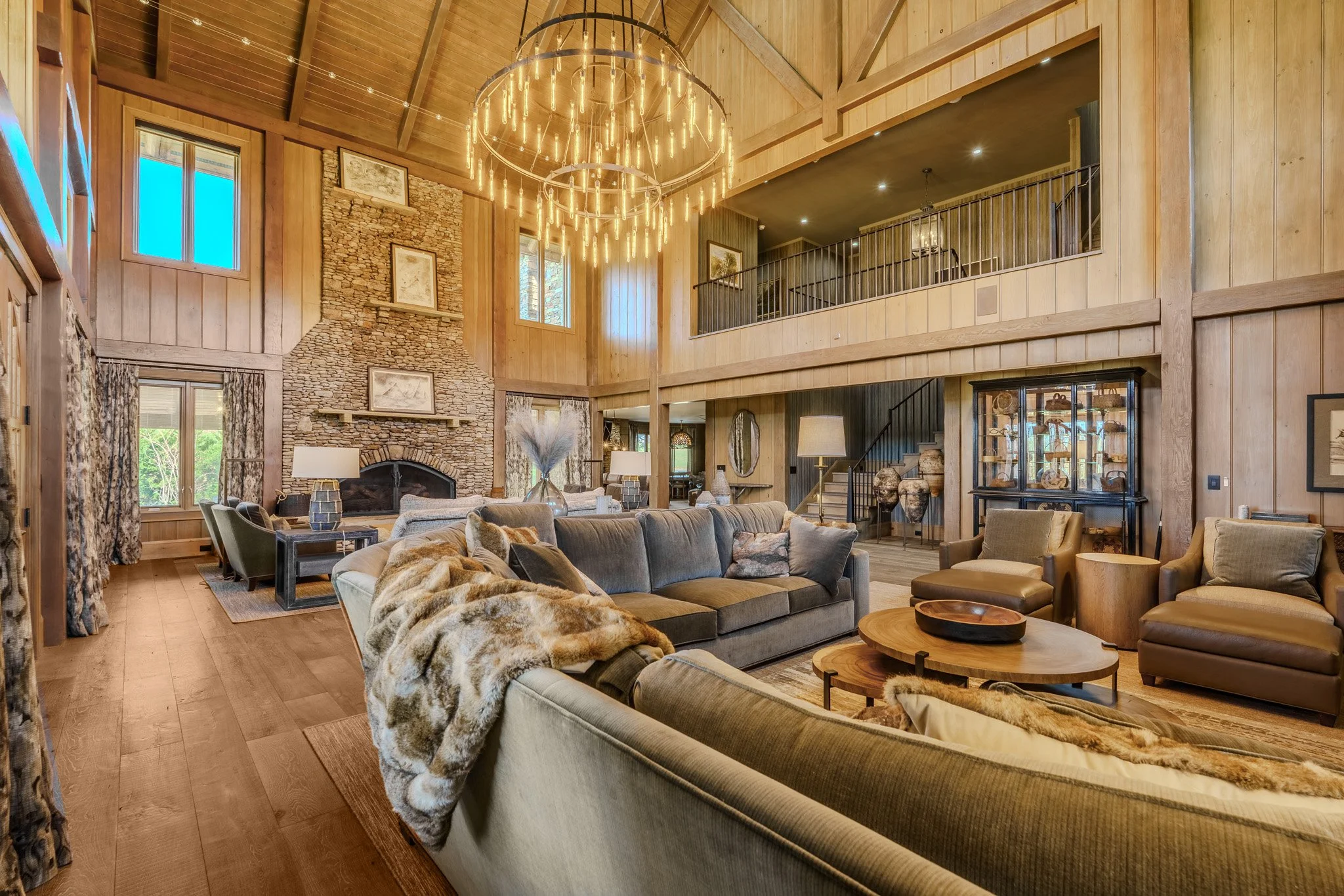 Spacious living room with wood-paneled walls and ceiling, large stone fireplace, multiple windows, modern chandelier, sectional sofa with throw blanket, and various chairs and tables.