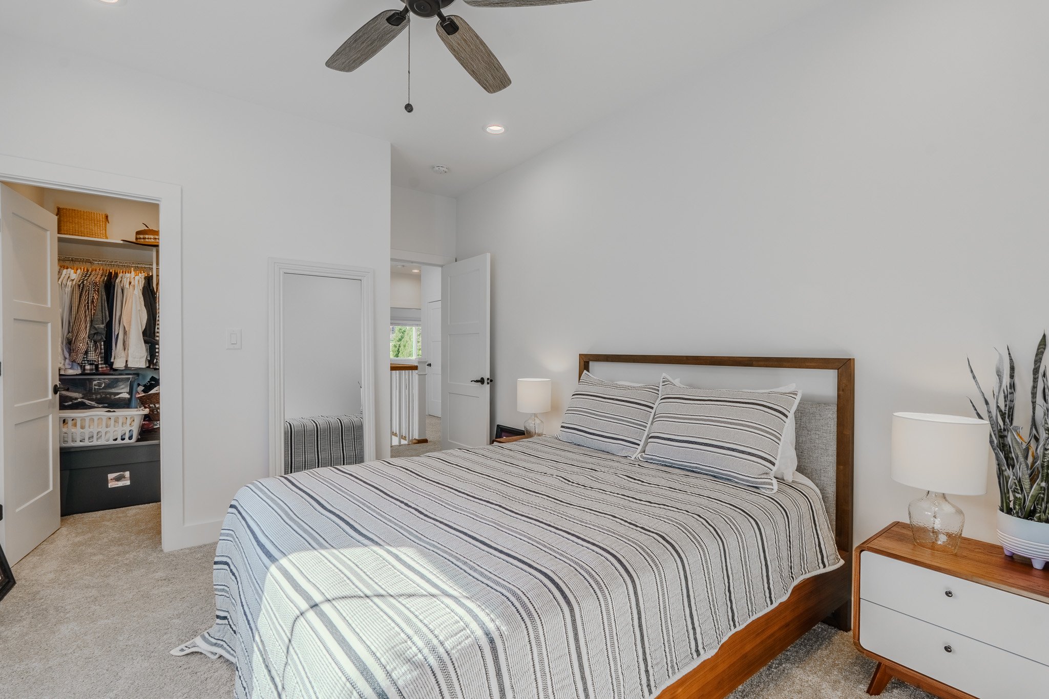 A bedroom with a bed covered in striped bedding, two matching pillows, nightstand with a white lamp and potted plant, open closet with clothes, and a ceiling fan.