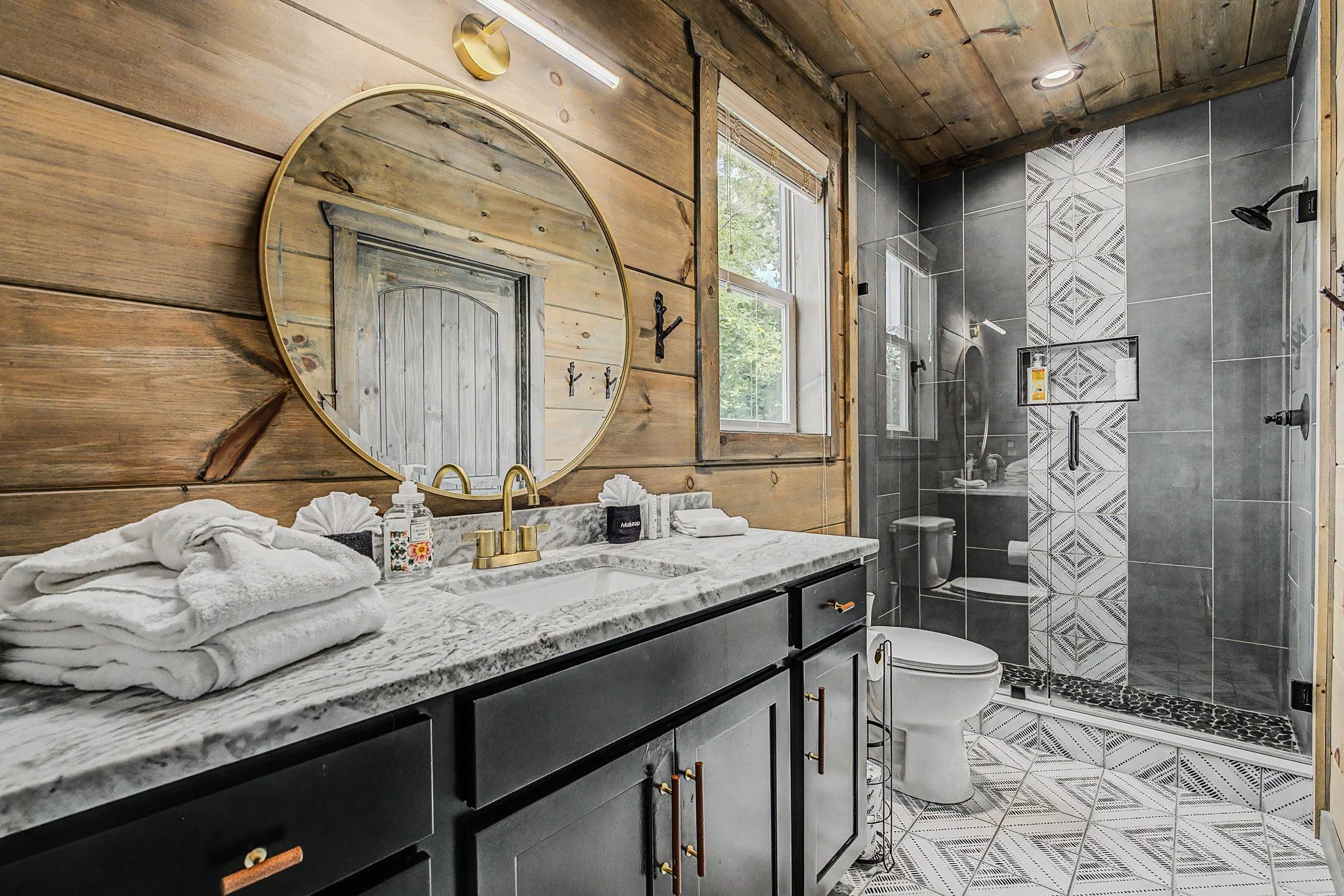 A bathroom featuring a wooden wall with a round mirror, a window with blinds, a marble countertop with a sink, and a shower area with patterned tiles and multiple showerheads.