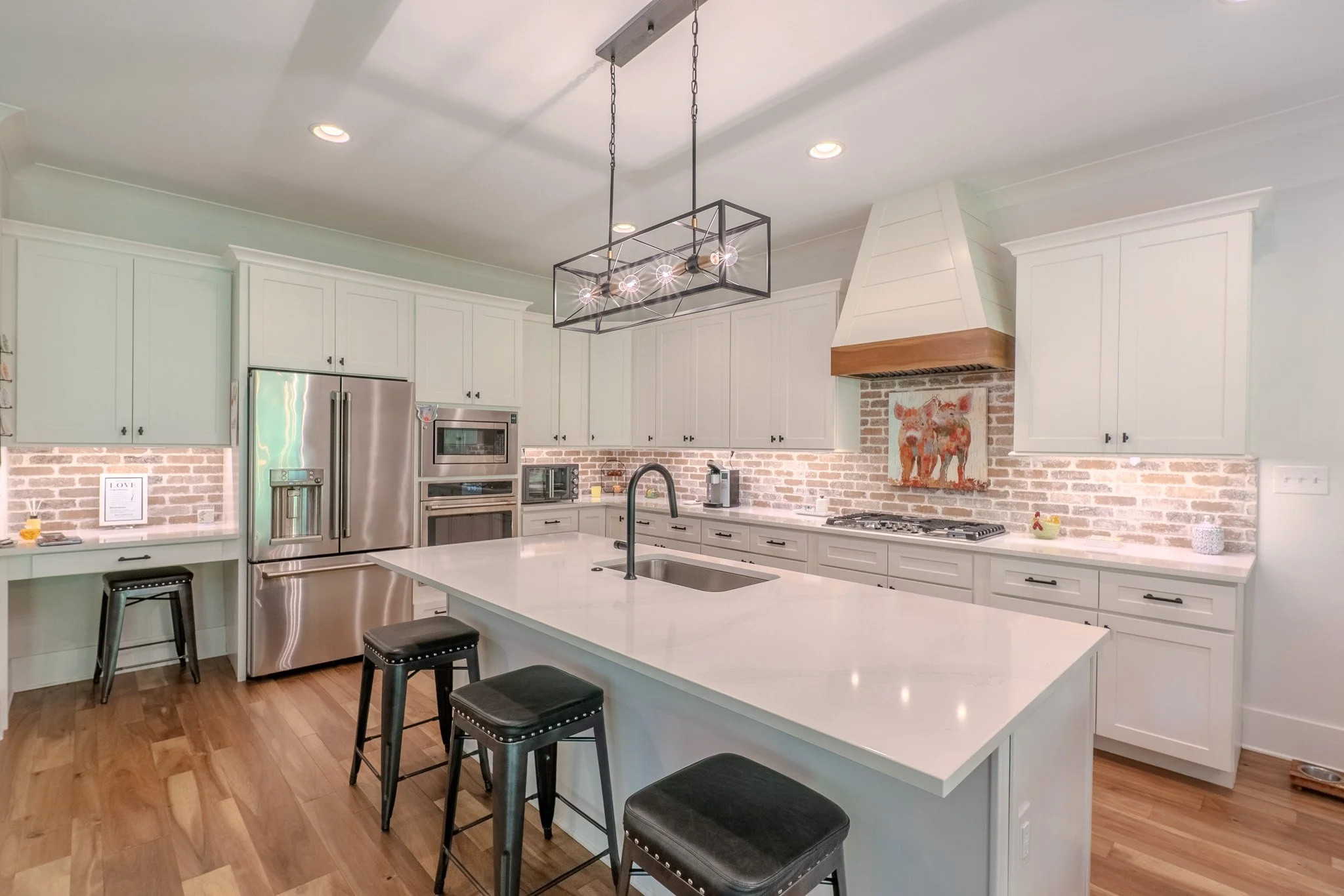 Modern kitchen with white cabinets, a large white island with a black faucet, stainless steel refrigerator, built-in oven and microwave, brick backsplash, and a wooden range hood above a stove, with pendant lighting hanging over the island.