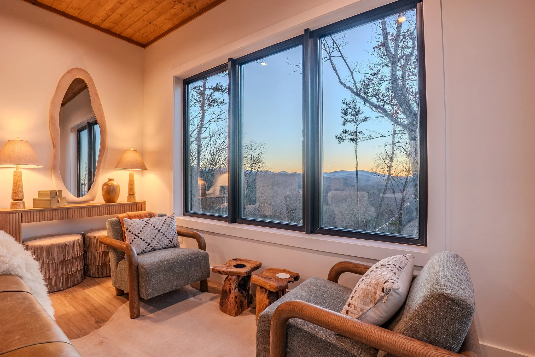 Living room with large window overlooking a scenic outdoor landscape with hills and trees at sunset, featuring two armchairs, a neutral-colored sofa, wooden side tables, and a wooden ceiling.