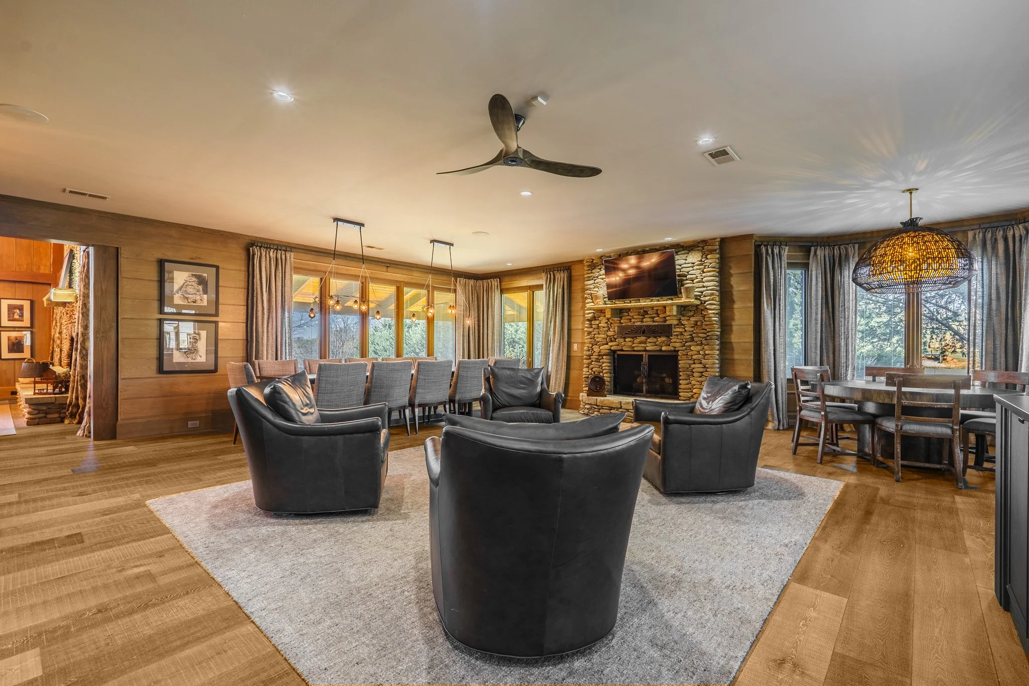 Living room with leather armchairs, a stone fireplace, large windows with curtains, a television, and a dining table with chairs.