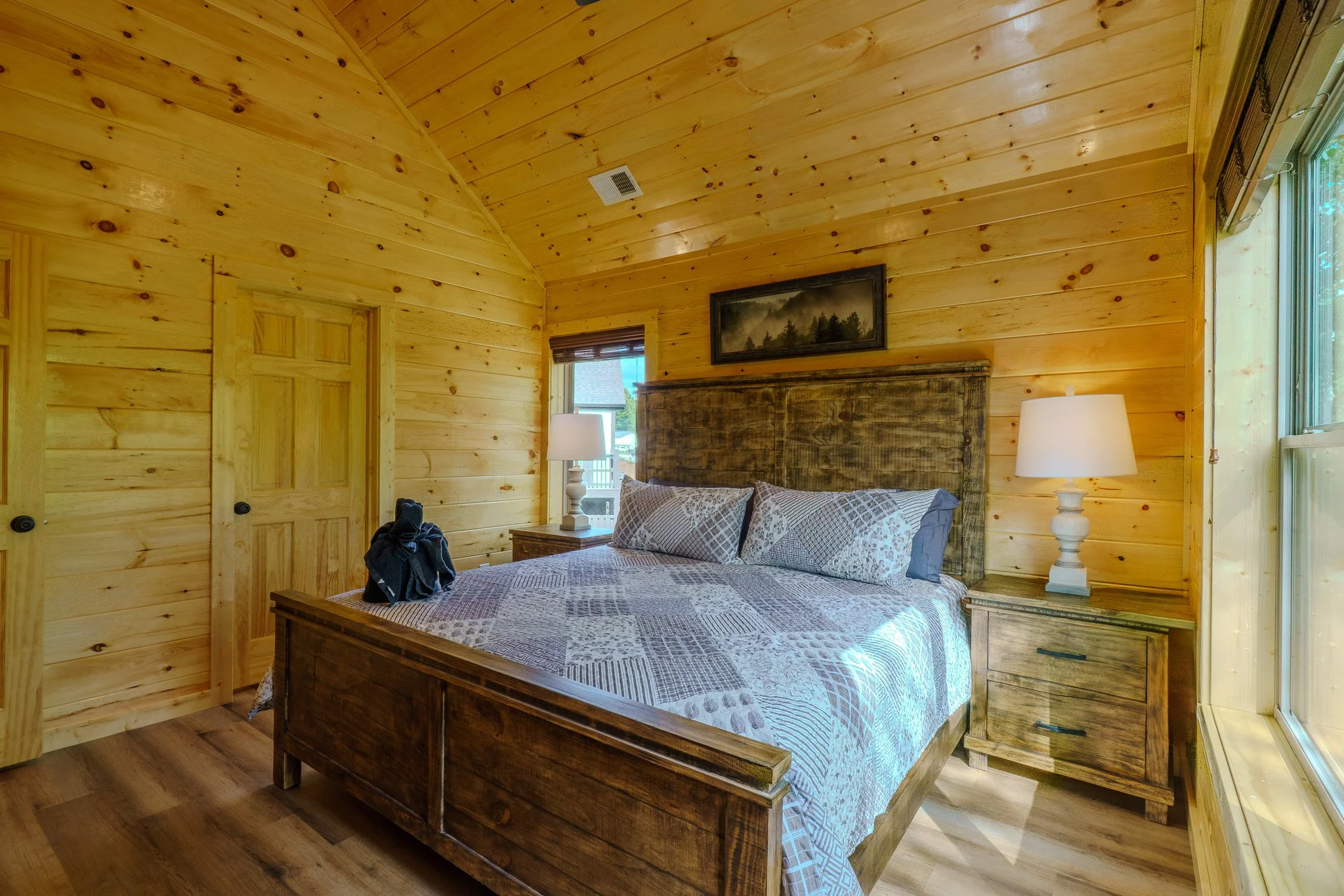 A bedroom with wooden walls, a large bed with patterned bedding, wooden nightstands with white lamps, a painting above the headboard, a window with natural light, and a wooden door.