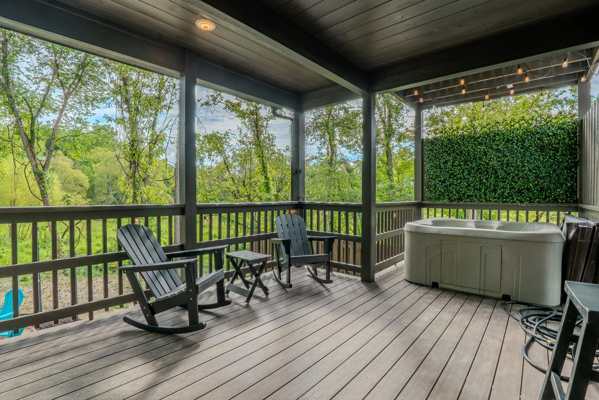 Covered porch with wooden flooring, two black Adirondack chairs, a small matching table, string lights hanging from ceiling, plastic hot tub, and a green hedge wall, overlooking green trees and sky.