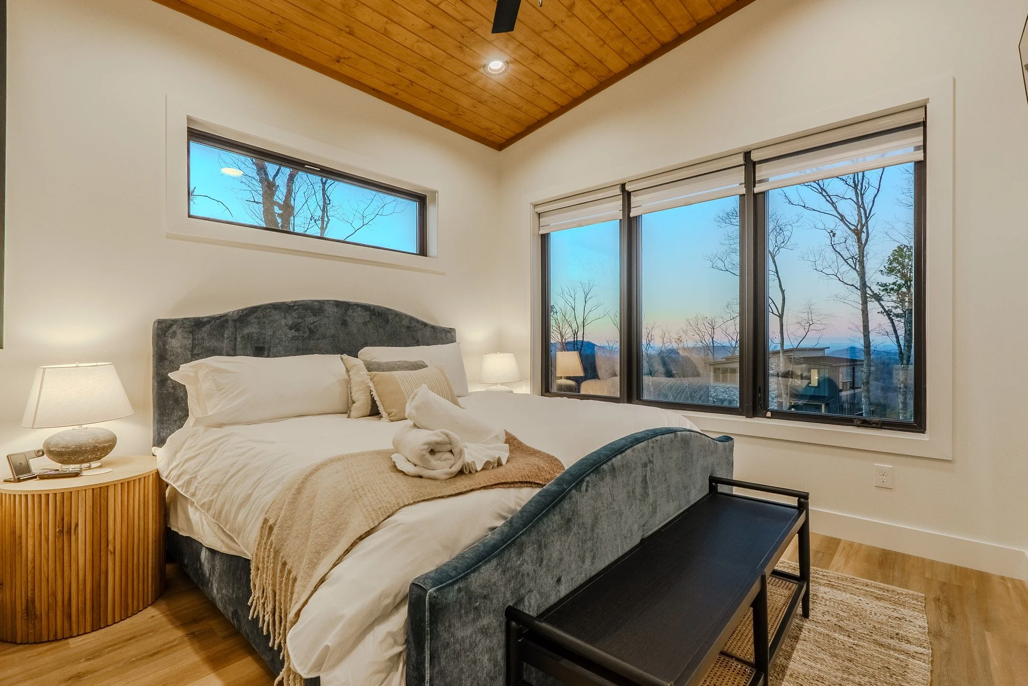 Bedroom with a large bed, rolled towels, nightstand with lamp, large windows showing trees and a sunset or sunrise scene, and wooden ceiling.