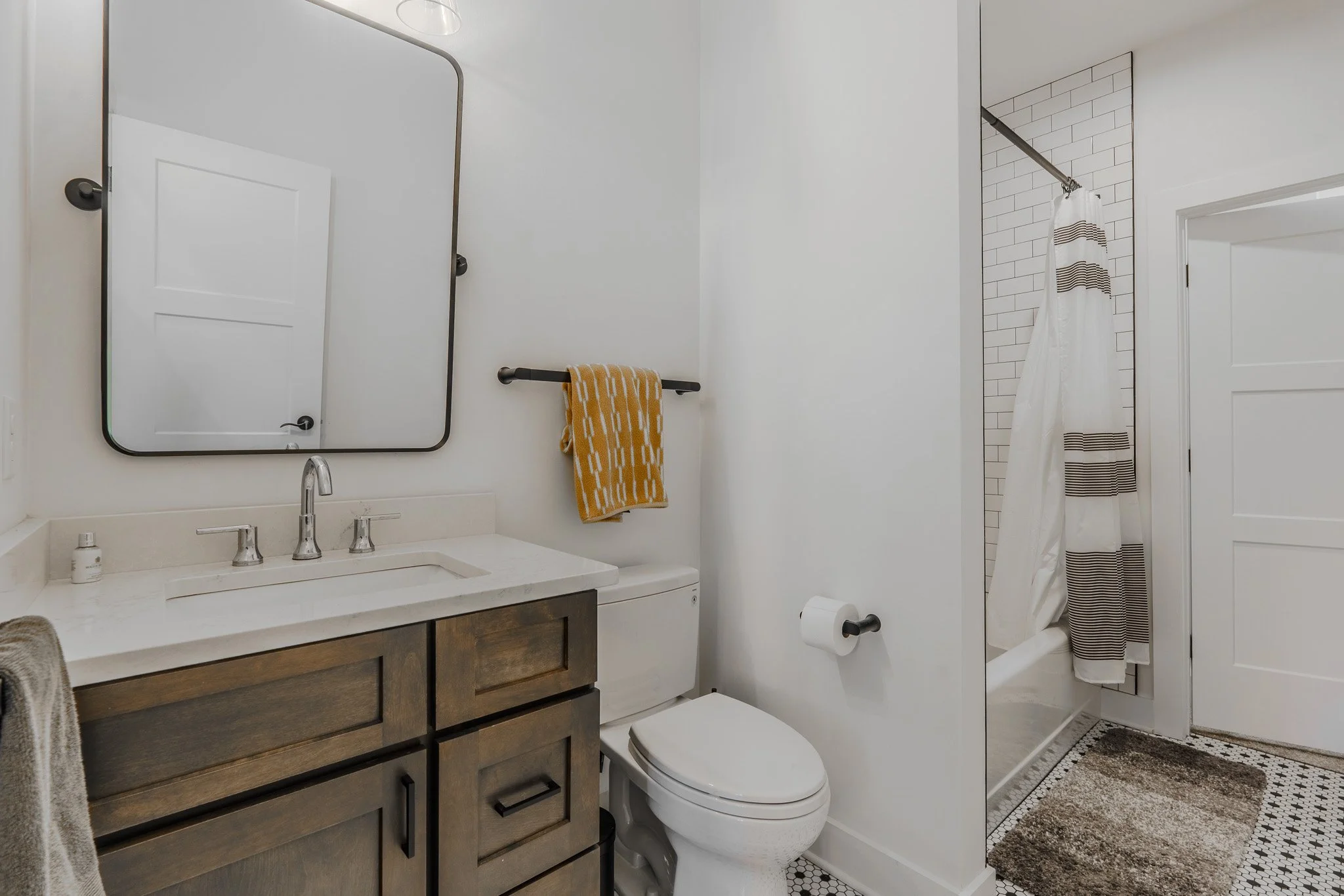 Bathroom with white walls, a vanity with dark wood cabinets, a white countertop, a mirror, a toilet with a yellow towel hanging above it, a toilet paper holder, a shower with a patterned curtain, and a backsplash of white subway tiles.