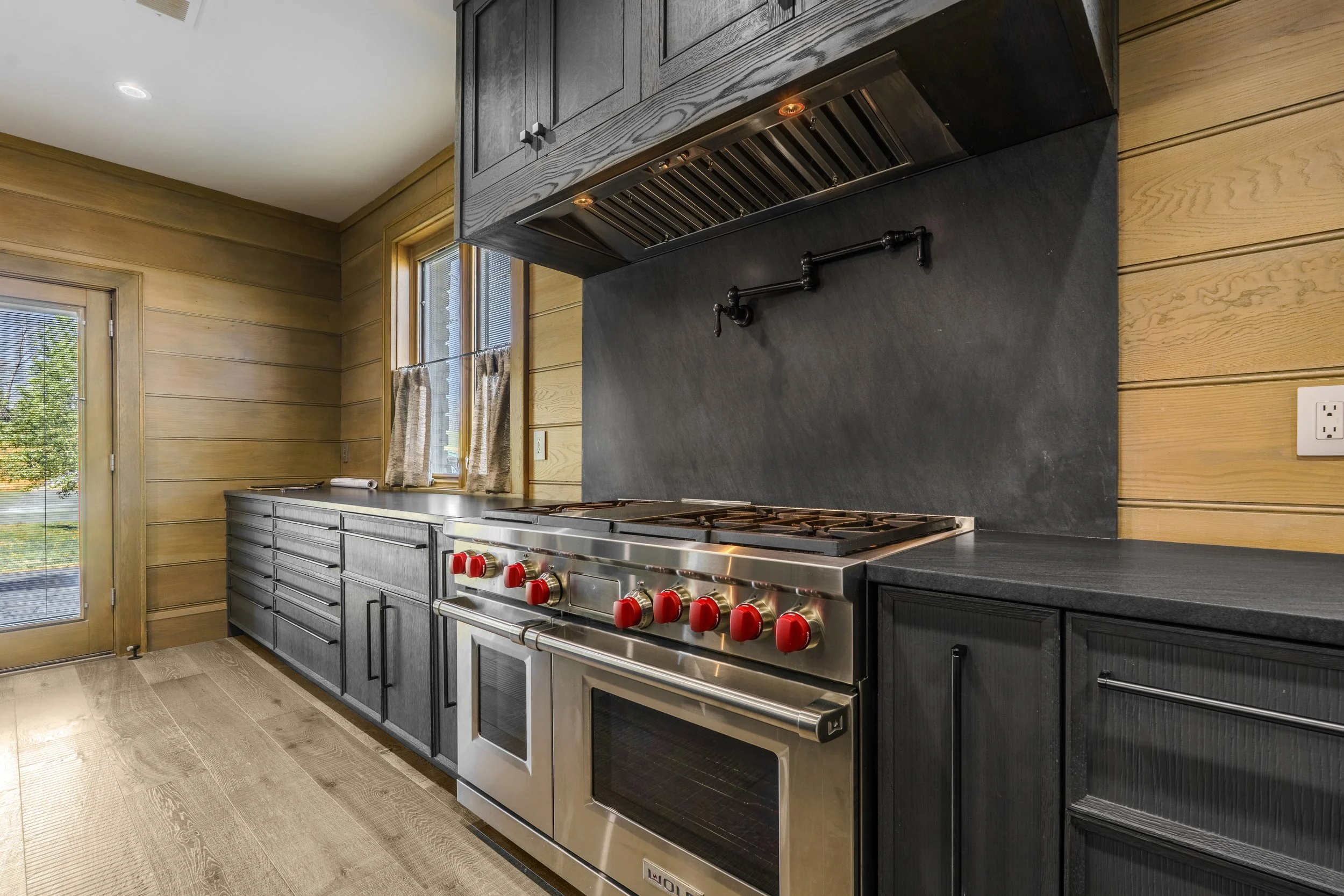 Modern kitchen with black cabinetry, stainless steel gas stove with red knobs, black backsplash, wooden walls, window with beige curtains, and a glass door leading outside.