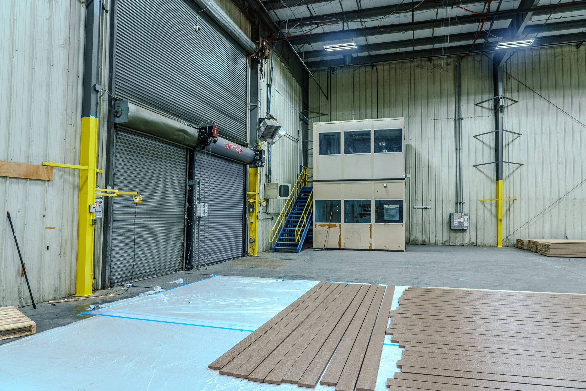 Interior of a warehouse with a large industrial roll-up door, a small elevated office area with stairs, wooden planks on a plastic sheet, and yellow safety markings.