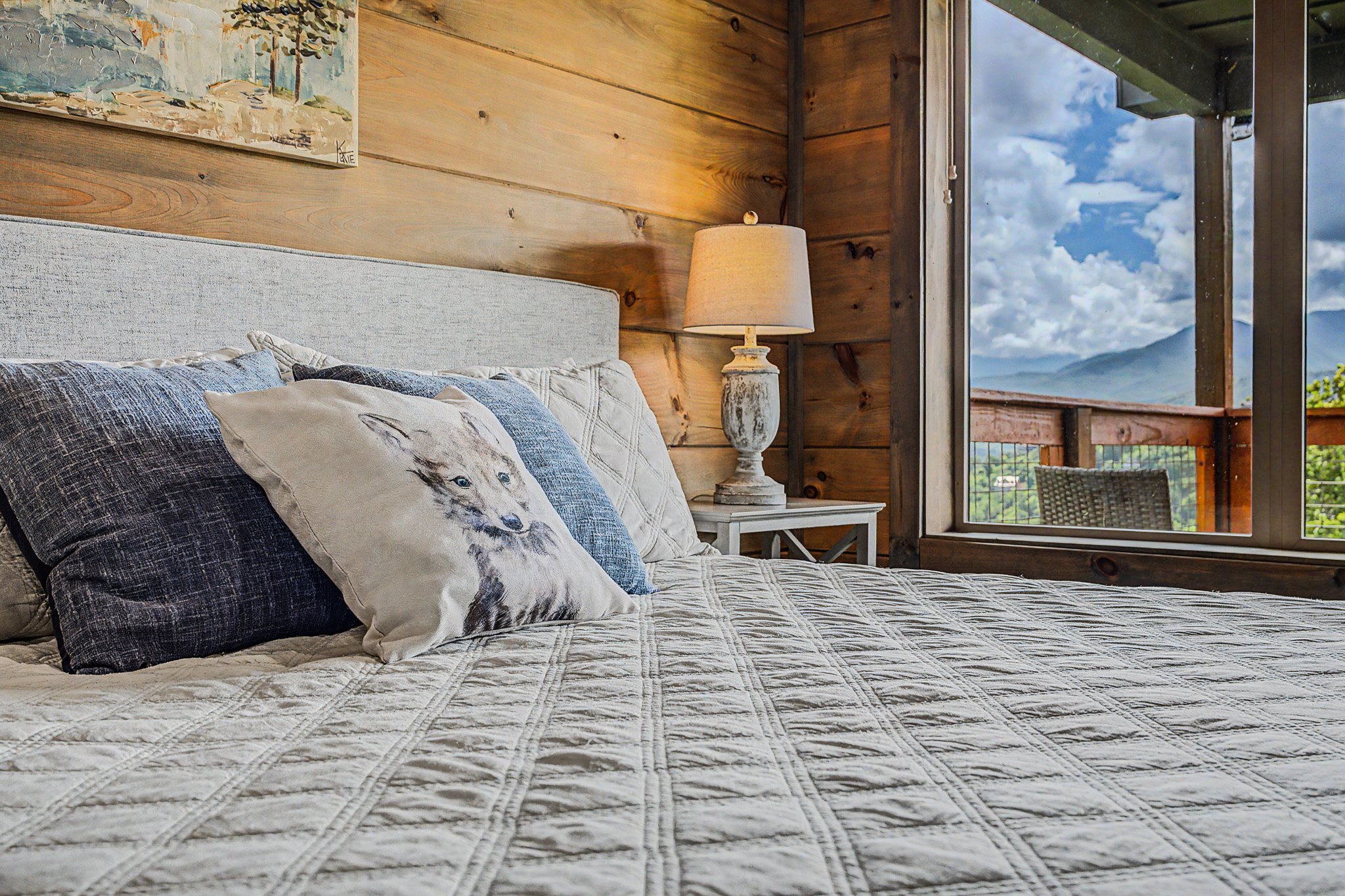 A cozy bedroom with a bed covered in a white quilt, decorated with pillows including one with a wolf face, beside a white bedside table with a rustic-looking lamp. The room features wooden walls and large windows opening to a mountain view and blue s