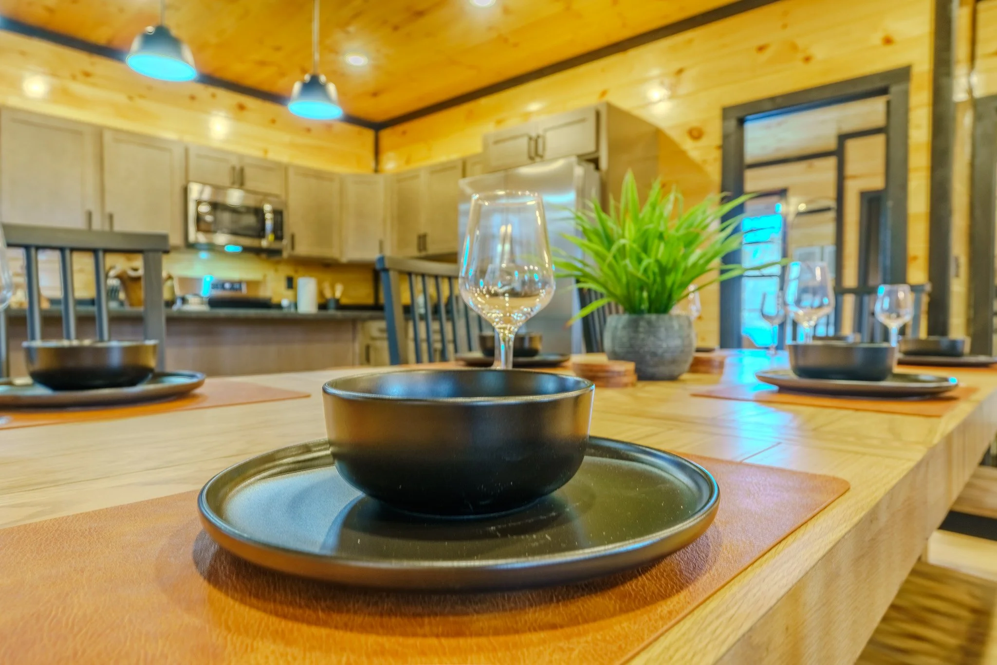 A dining table set with black bowls, glasses, and placemats, in a cozy wooden kitchen with a potted green plant, pendant lights, and a large refrigerator.