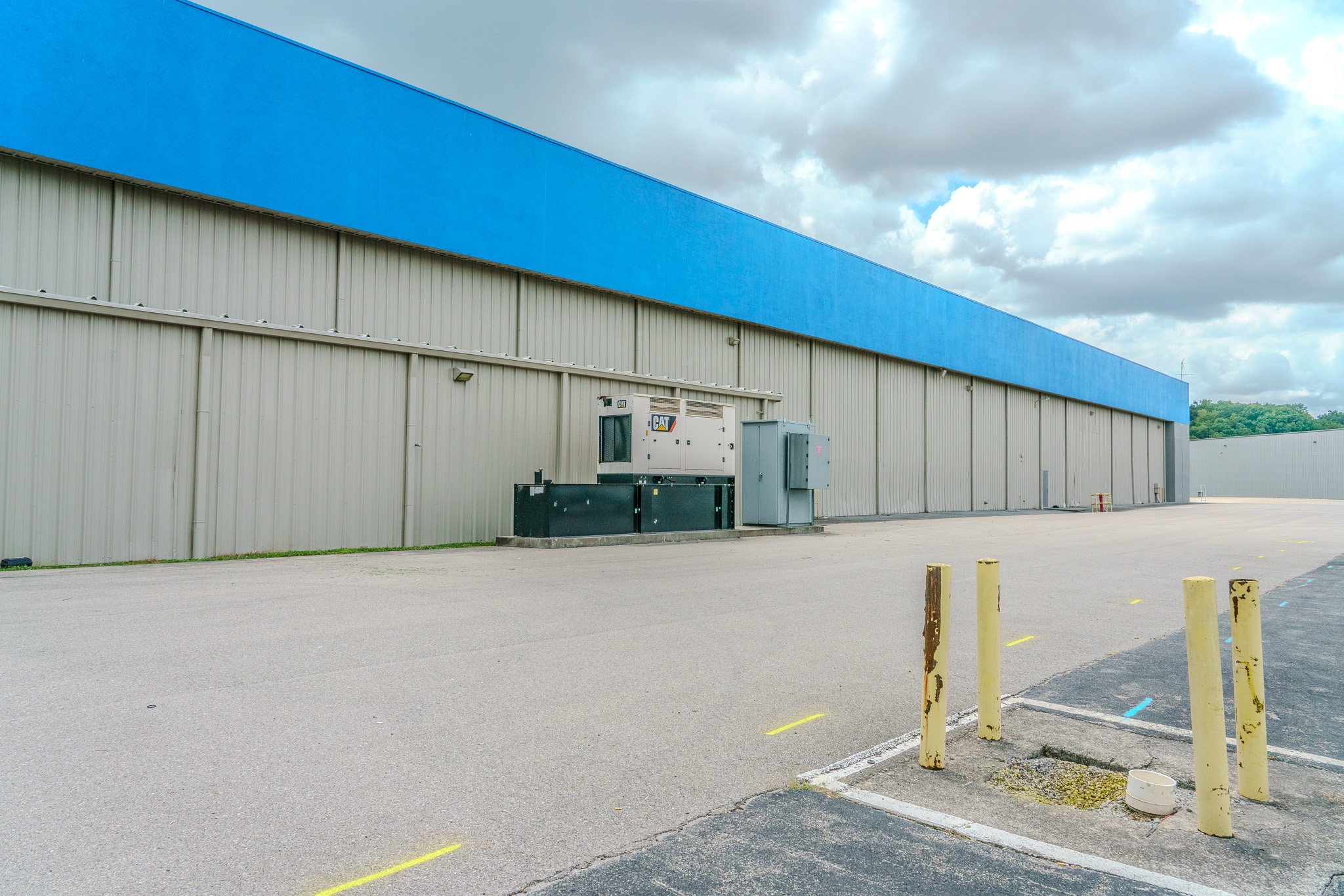 An empty parking lot with a large industrial building featuring beige metal siding and a blue awning, under a partly cloudy sky.