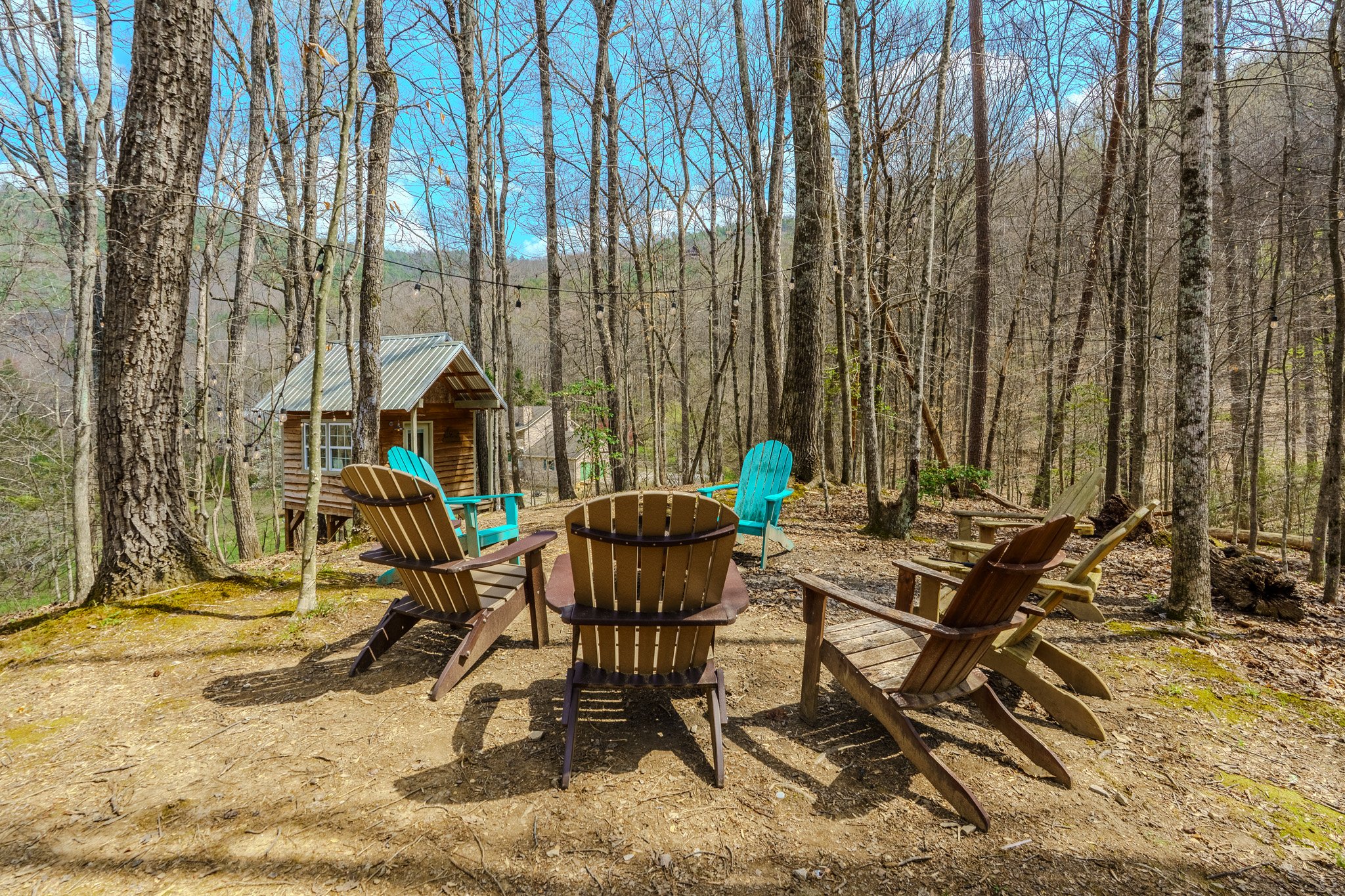 A cozy outdoor seating area with Adirondack chairs in various shades of brown, turquoise, and green, situated in a wooded forest with a small wooden cabin in the background and leafless trees around.
