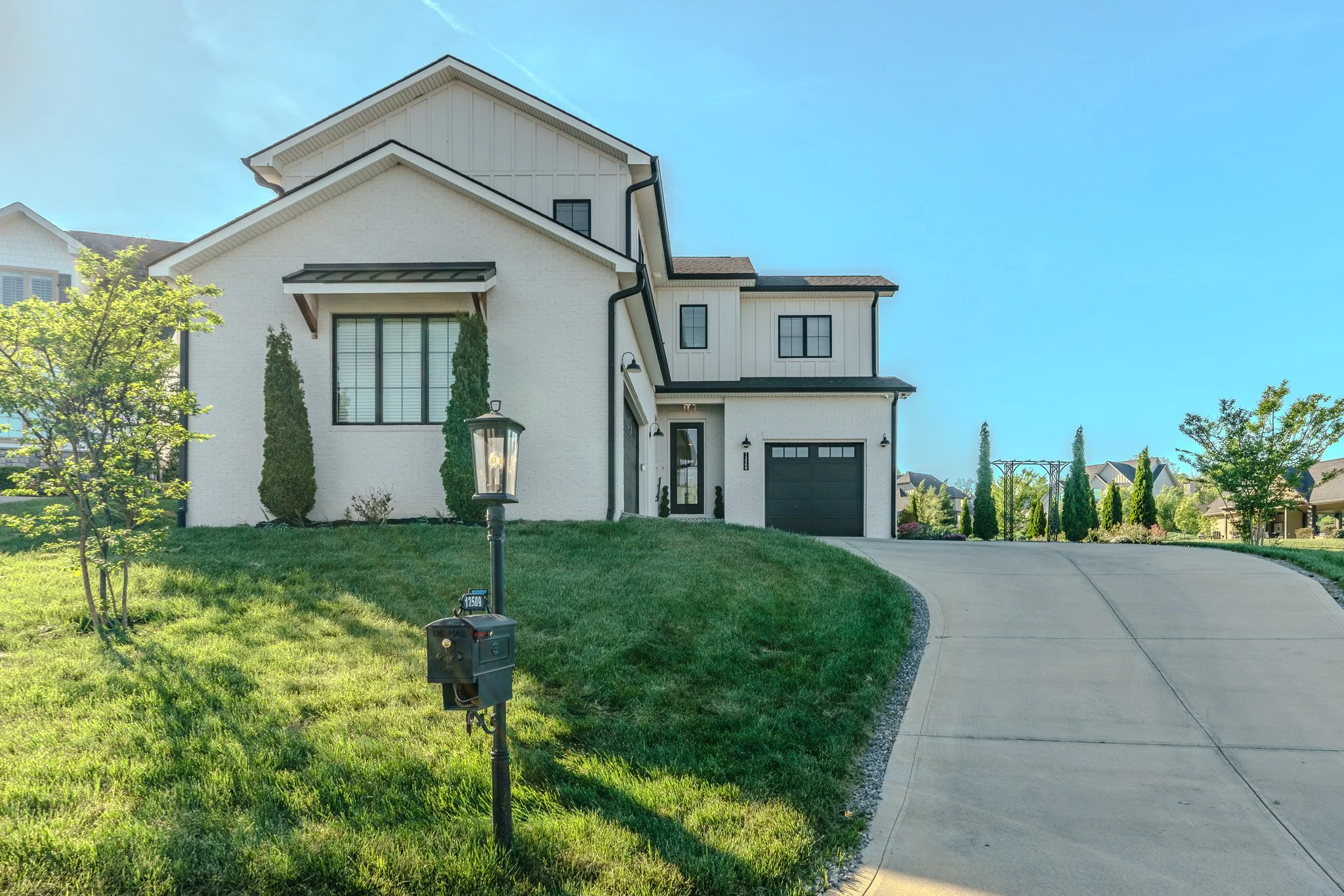Modern two-story white house with black garage door, driveway, front lawn, trees, and a lamp post with mailbox.
