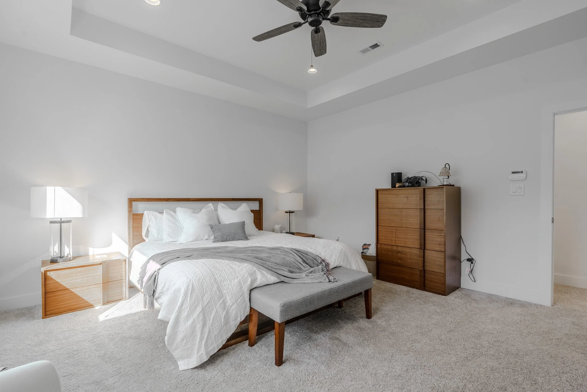 A bedroom with a large bed with white bedding and pillows, two wooden nightstands with lamps, a gray upholstered bench at the foot of the bed, a wooden dresser with various electronic items on top, and a ceiling fan with dark blades.