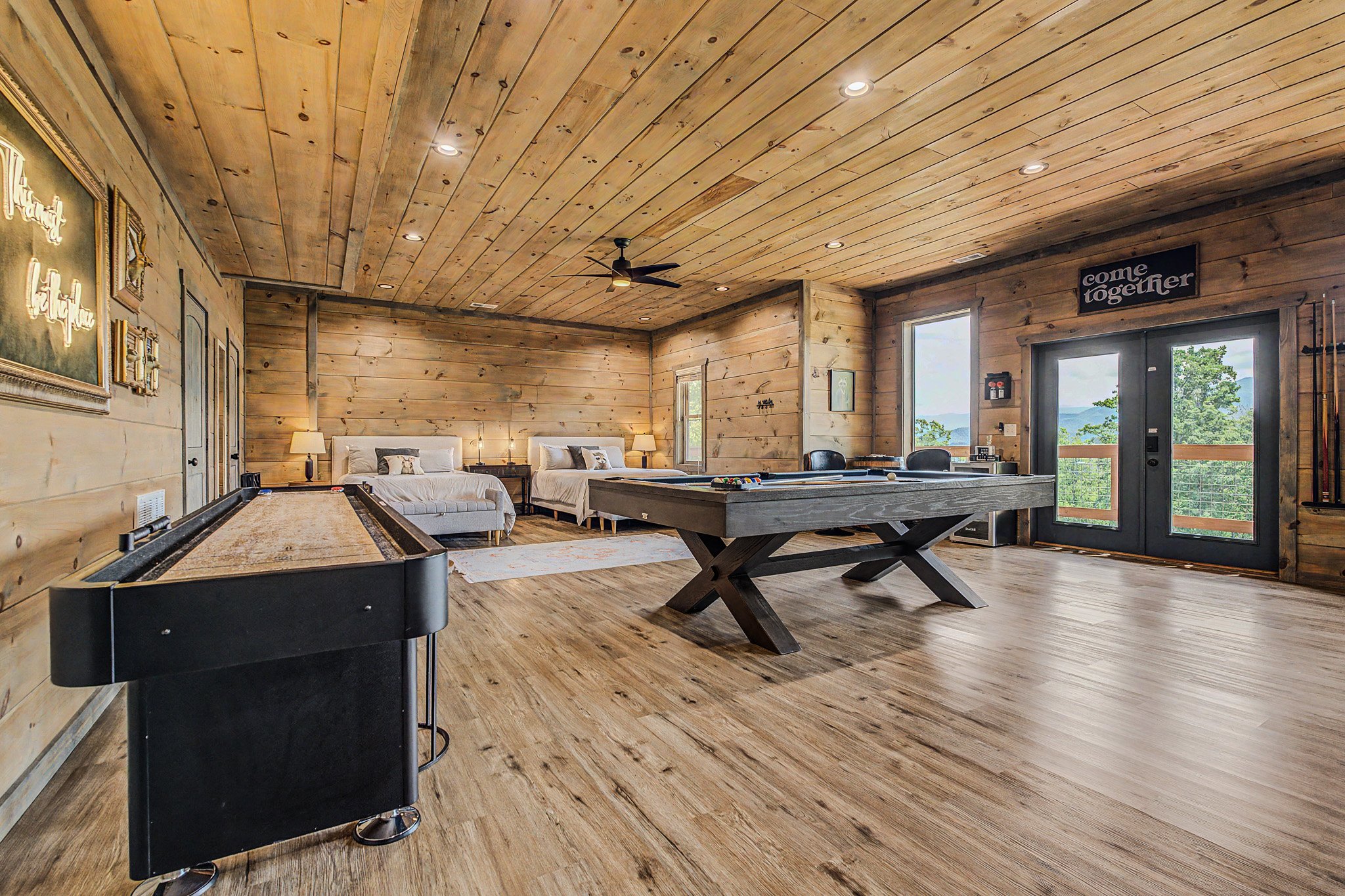 A spacious recreational room with wooden walls, ceiling, and floor. It features a pool table, two beds with bedside lamps, a foosball table, wall art, a ceiling fan, large windows, and a sliding glass door showing a view of trees and mountains outsid