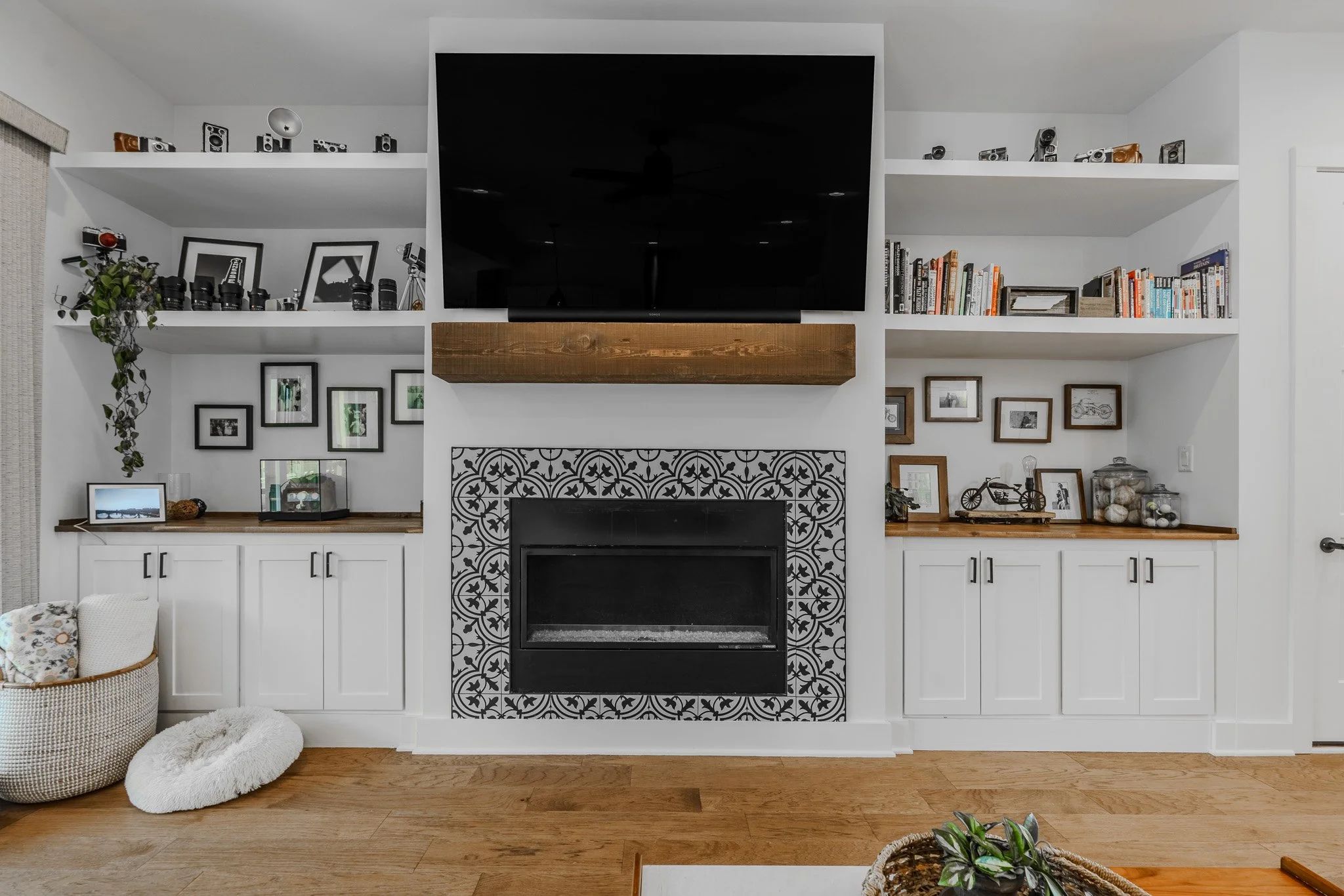 Living room with a fireplace, TV mounted above the fireplace, built-in white shelves on both sides filled with books, photos, and decorations, hardwood floors, and a basket with a blanket on the left.