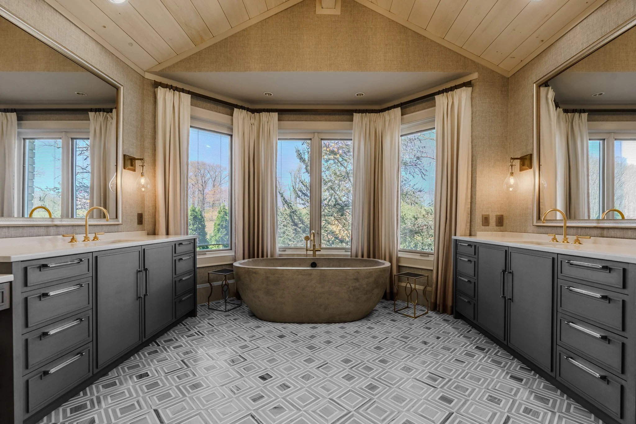 A spacious bathroom with a large windowed alcove featuring a freestanding bathtub. The room has two vanities with gray cabinets, white countertops, and gold fixtures. Large mirrors above each vanity, gold sconces, and patterned tile flooring.