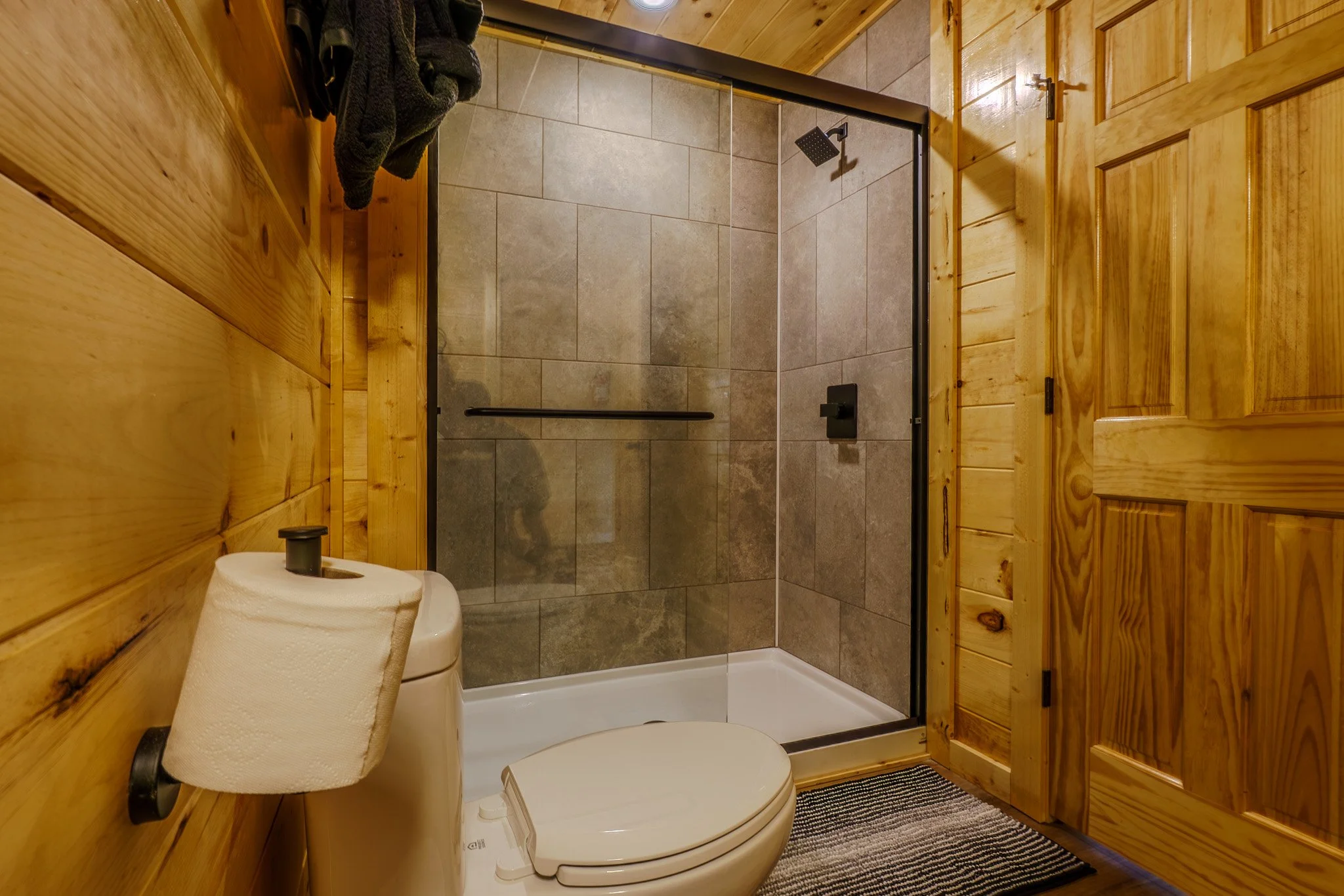 Bathroom with wooden walls, a toilet with a roll of toilet paper, and a glass-enclosed shower with tiled walls and a black shower fixture.