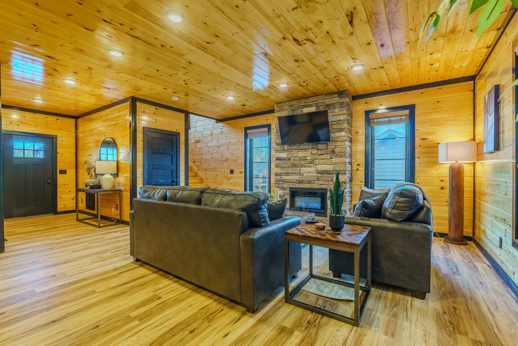 Living room with wooden walls and ceiling, black door, black-framed windows, a stone fireplace with a mounted TV, black leather couches, a wooden coffee table, potted plant, lamp, floor lamp, and decorative items.