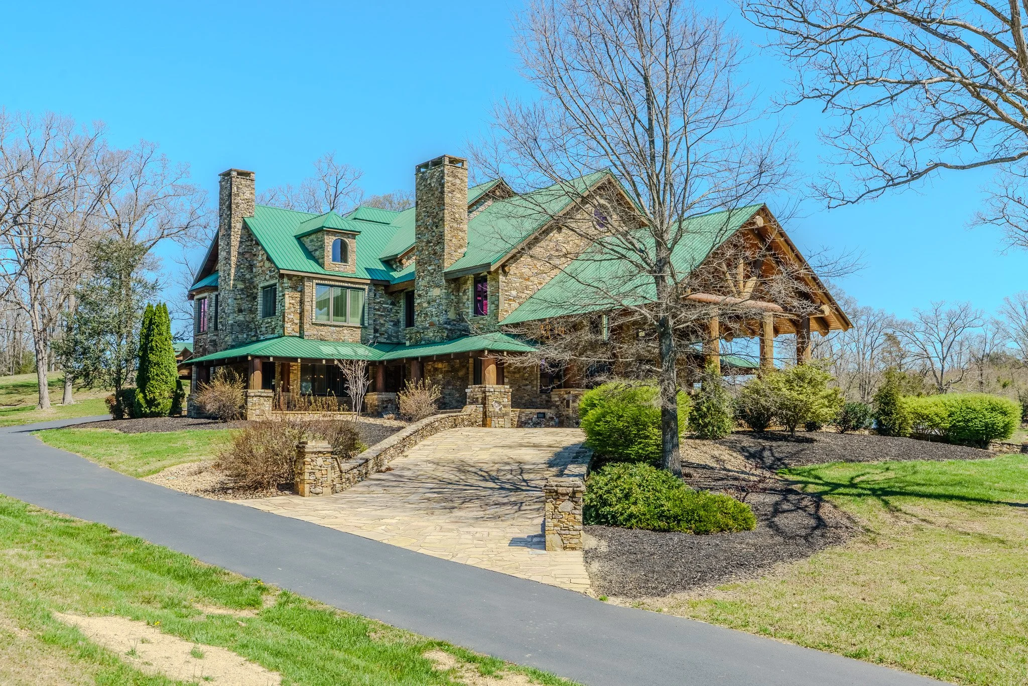 Large stone house with green metal roof, multiple chimneys, and large windows, surrounded by a landscaped yard with trees, shrubs, and a paved driveway.