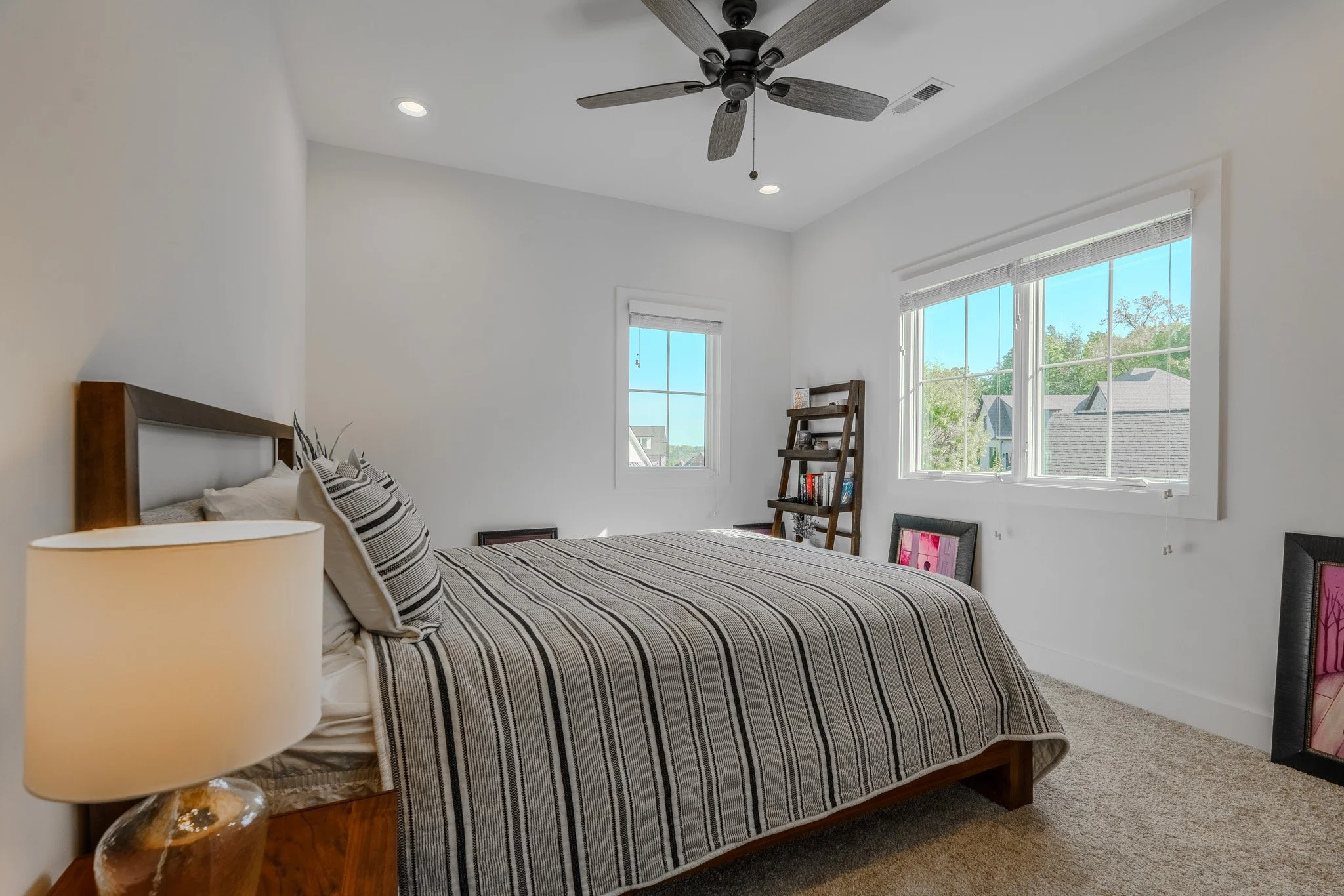 A minimalist bedroom with white walls, a wooden bed with striped bedding, a nightstand with a beige lamp, a ladder shelf with books, and large windows showing a suburban neighborhood outside.