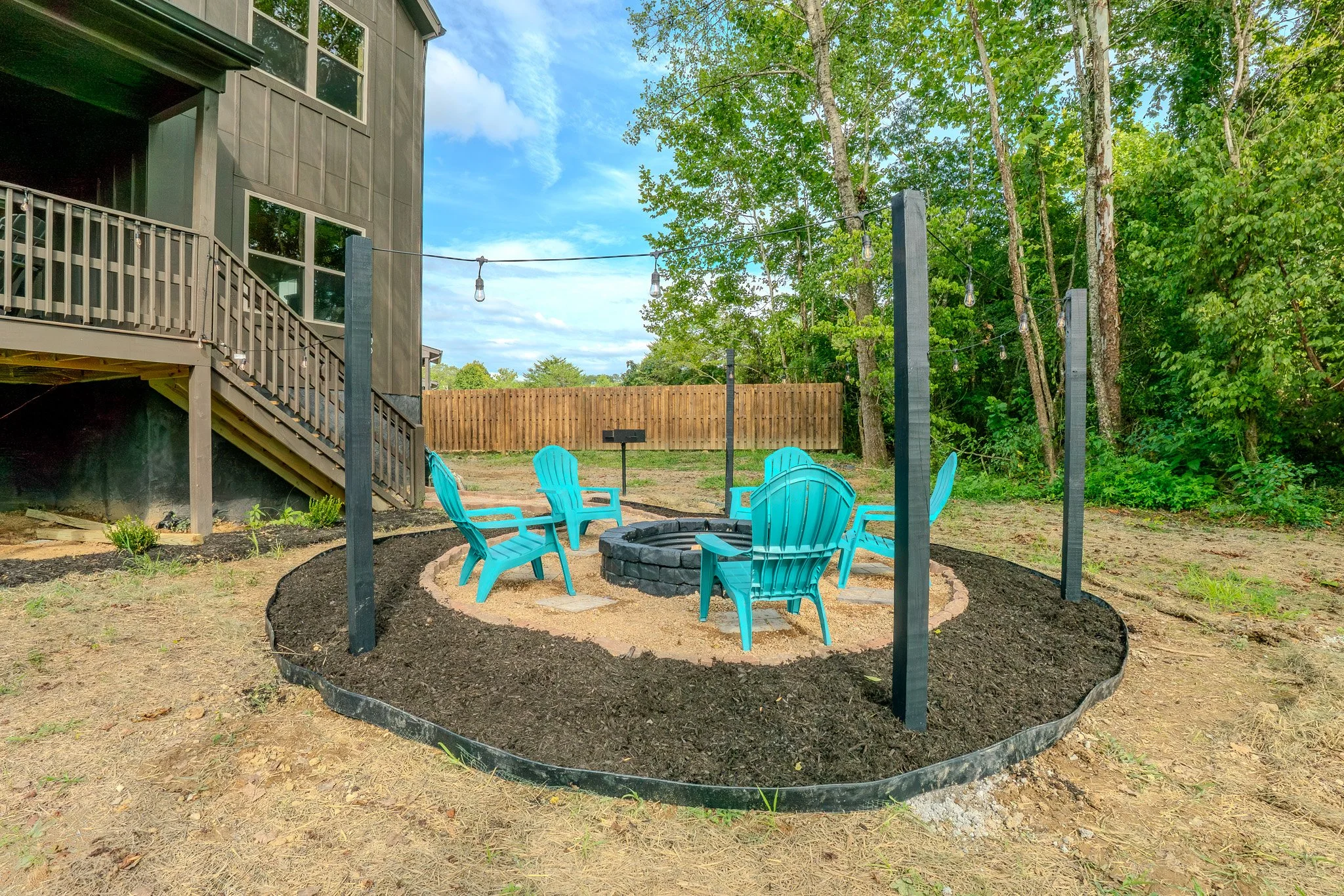 Outdoor backyard with a fire pit surrounded by turquoise Adirondack chairs, string lights overhead, and a wooden fence with trees in the background.