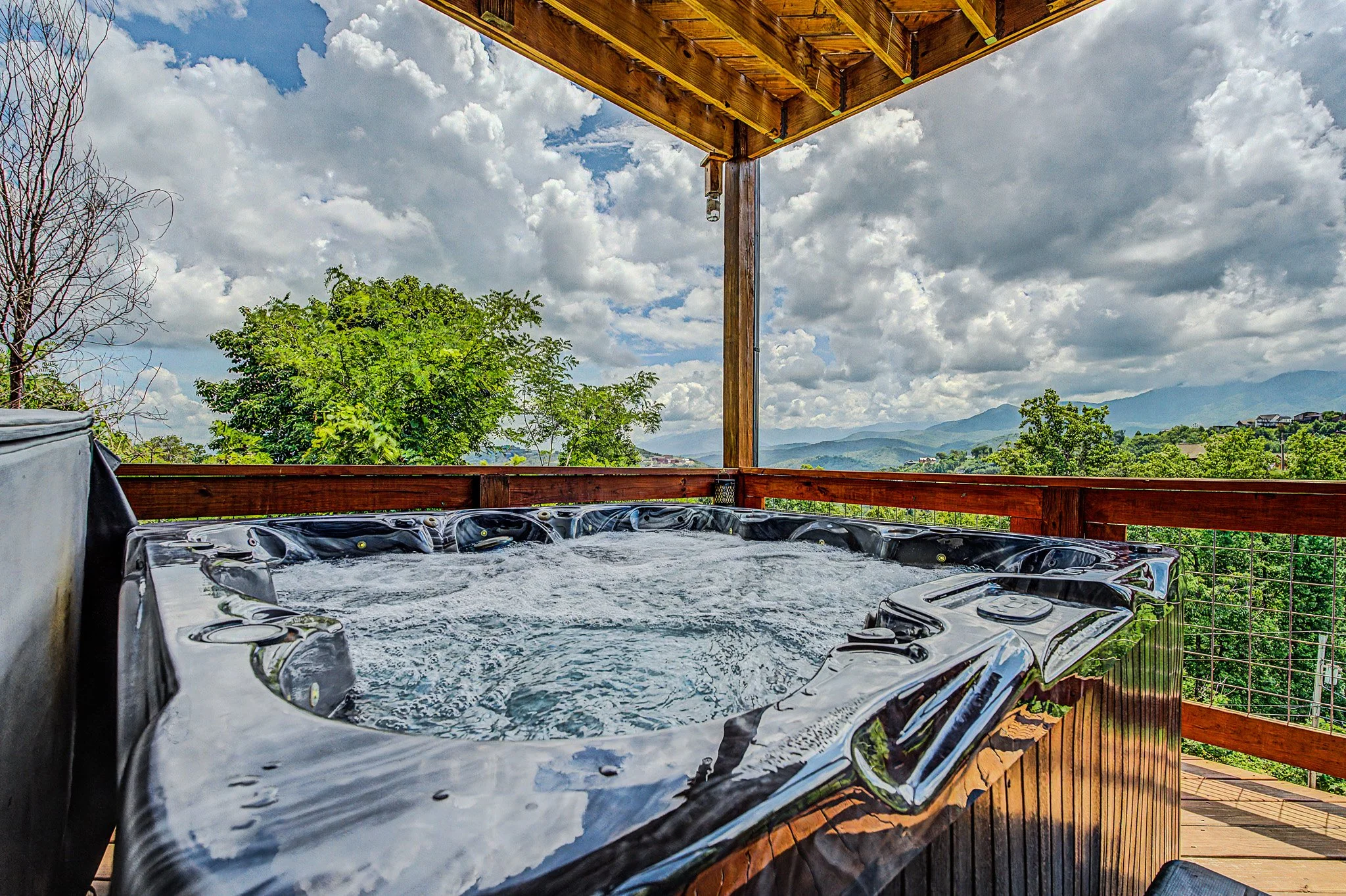 Outdoor wooden deck with hot tub filled with bubbling water, mountains and cloudy sky in the background.