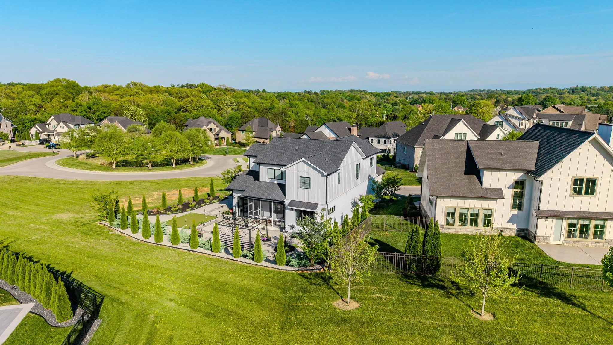 Aerial view of a residential neighborhood with modern houses, green lawns, trees, and a circular road in the background on a sunny day.