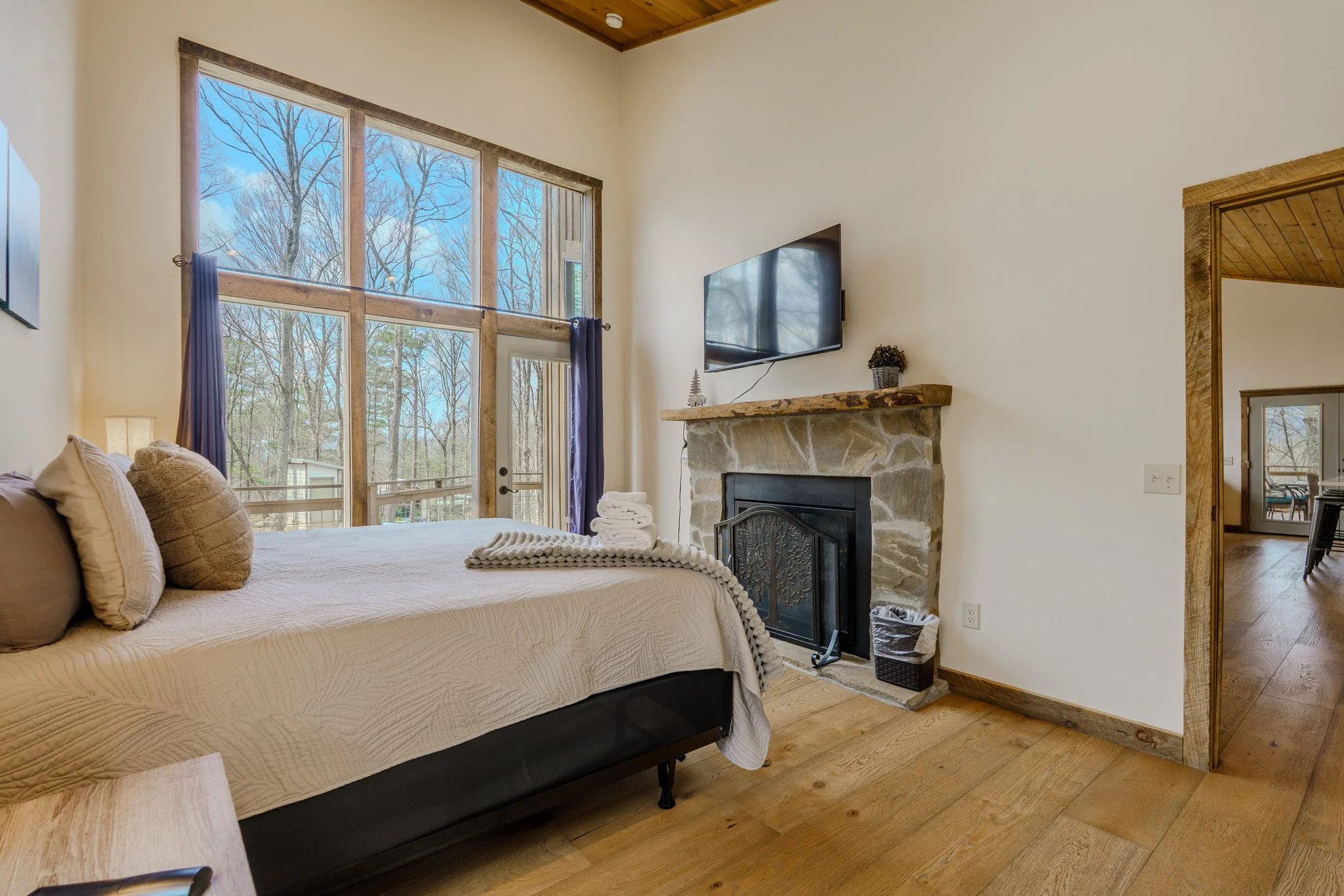 A cozy bedroom with a large window showing bare trees outside, a bed with beige pillows and a blanket, a stone fireplace with a wooden mantel, a mounted TV above the fireplace, and a door leading to a balcony or outdoor area.